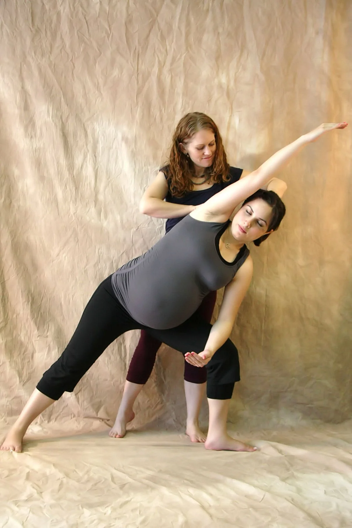 Two women practicing yoga against a brown textured background. One woman, pregnant, leans sideways with her right arm extended upward and her left arm resting on her thigh. The other woman supports her from behind.
