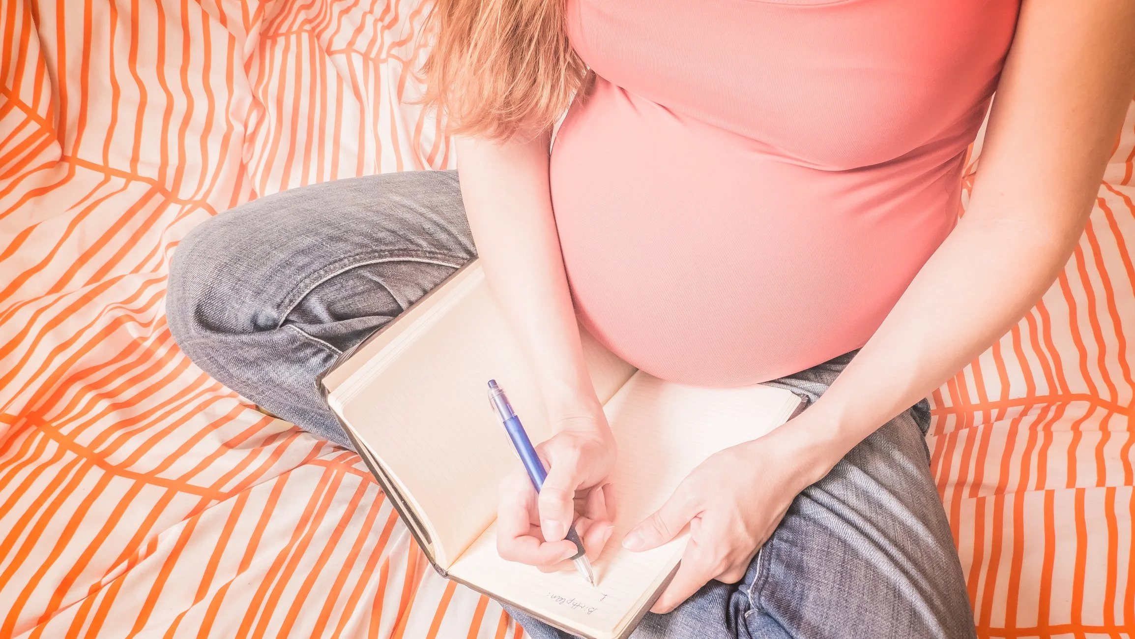 Pregnant woman sitting cross-legged on an orange striped bed, writing in a notebook with a blue pen.