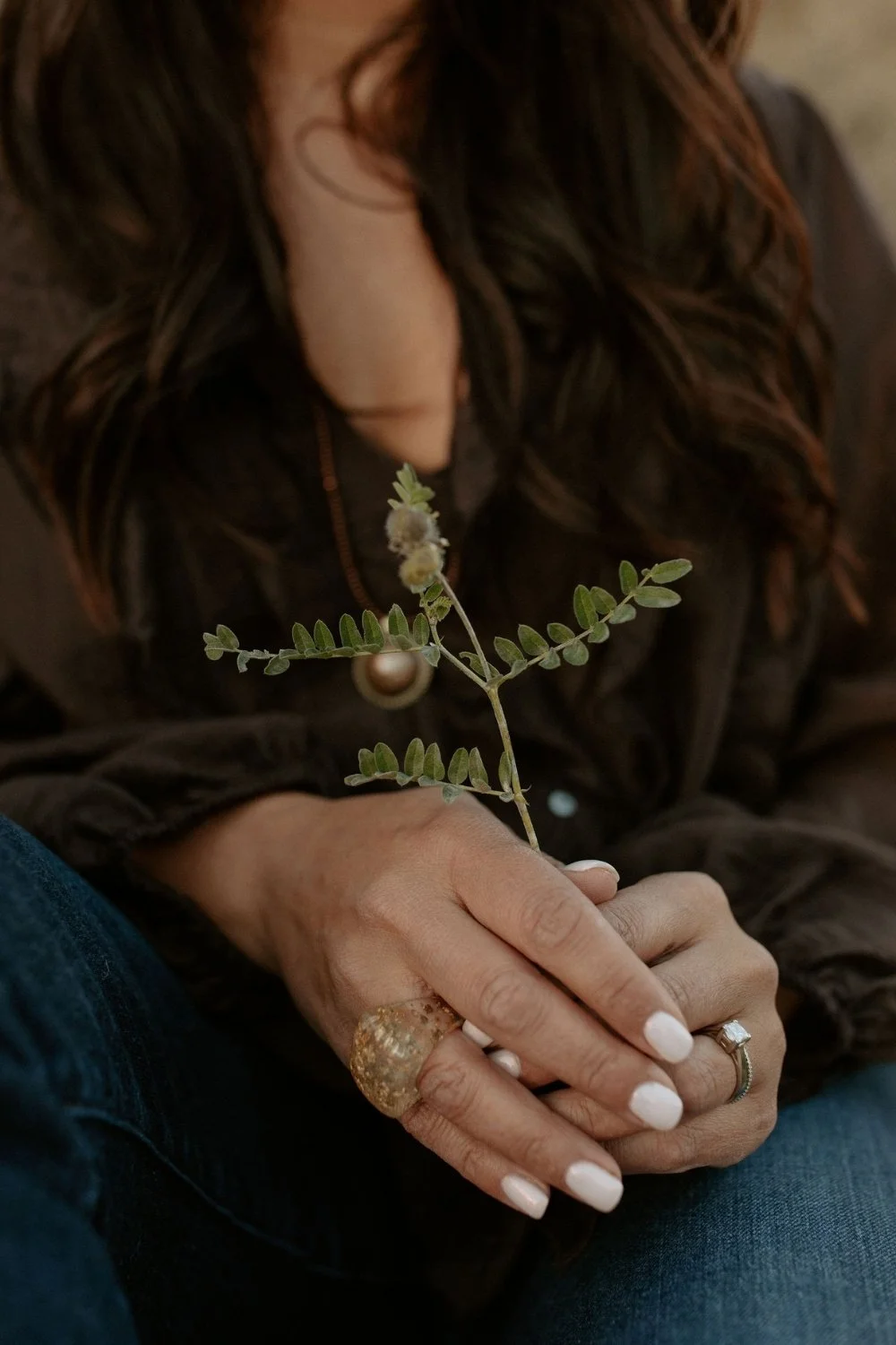 Close-up of a woman with wavy brown hair holding a small leafy plant with tiny flowers. She wears a large ring on her middle finger and a smaller ring on her ring finger, with her nails painted white. She is wearing a dark-colored jacket.