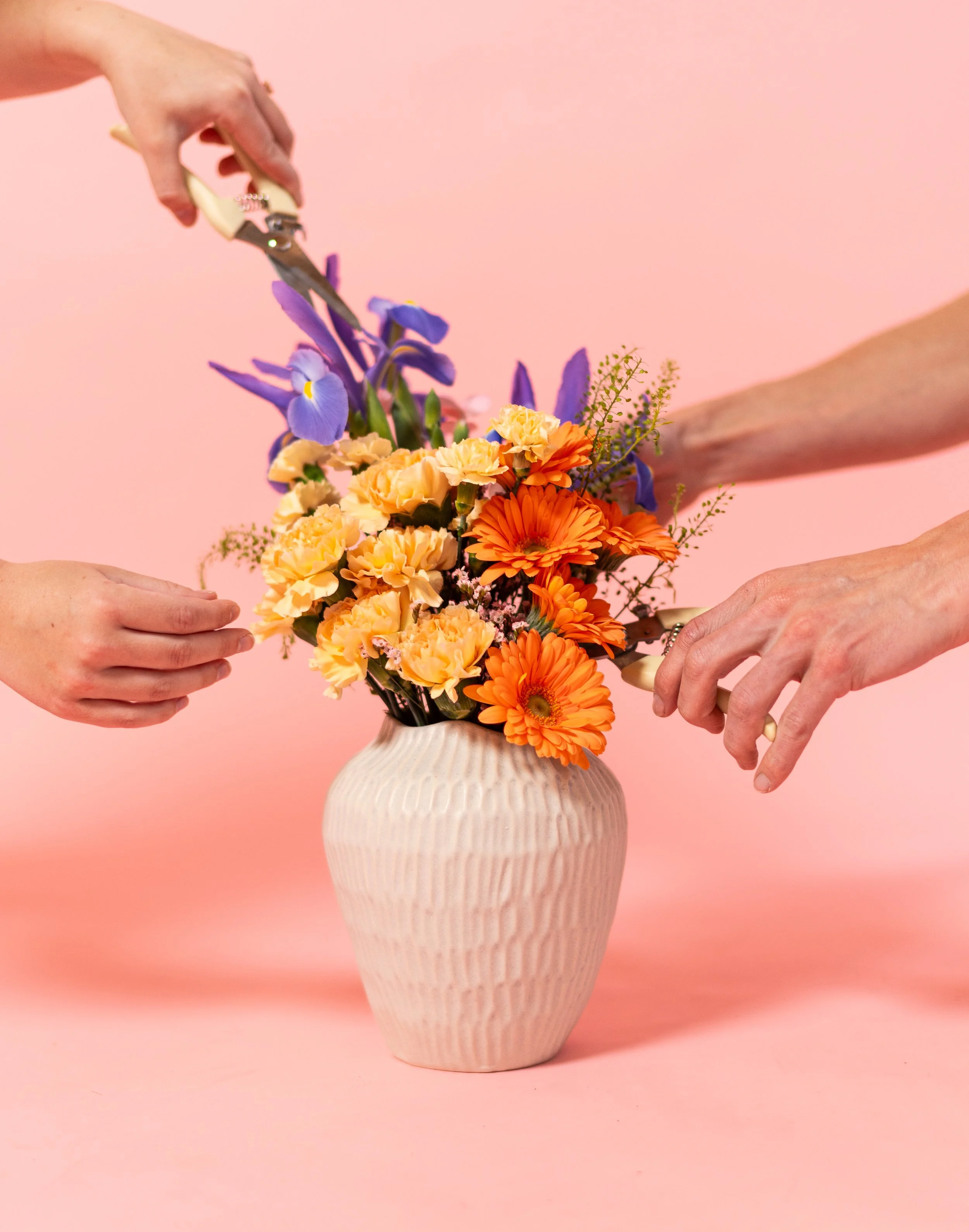 Several hands are arranging a bouquet of purple irises, orange gerbera daisies, and yellow carnations in a beige textured ceramic vase against a pink background.