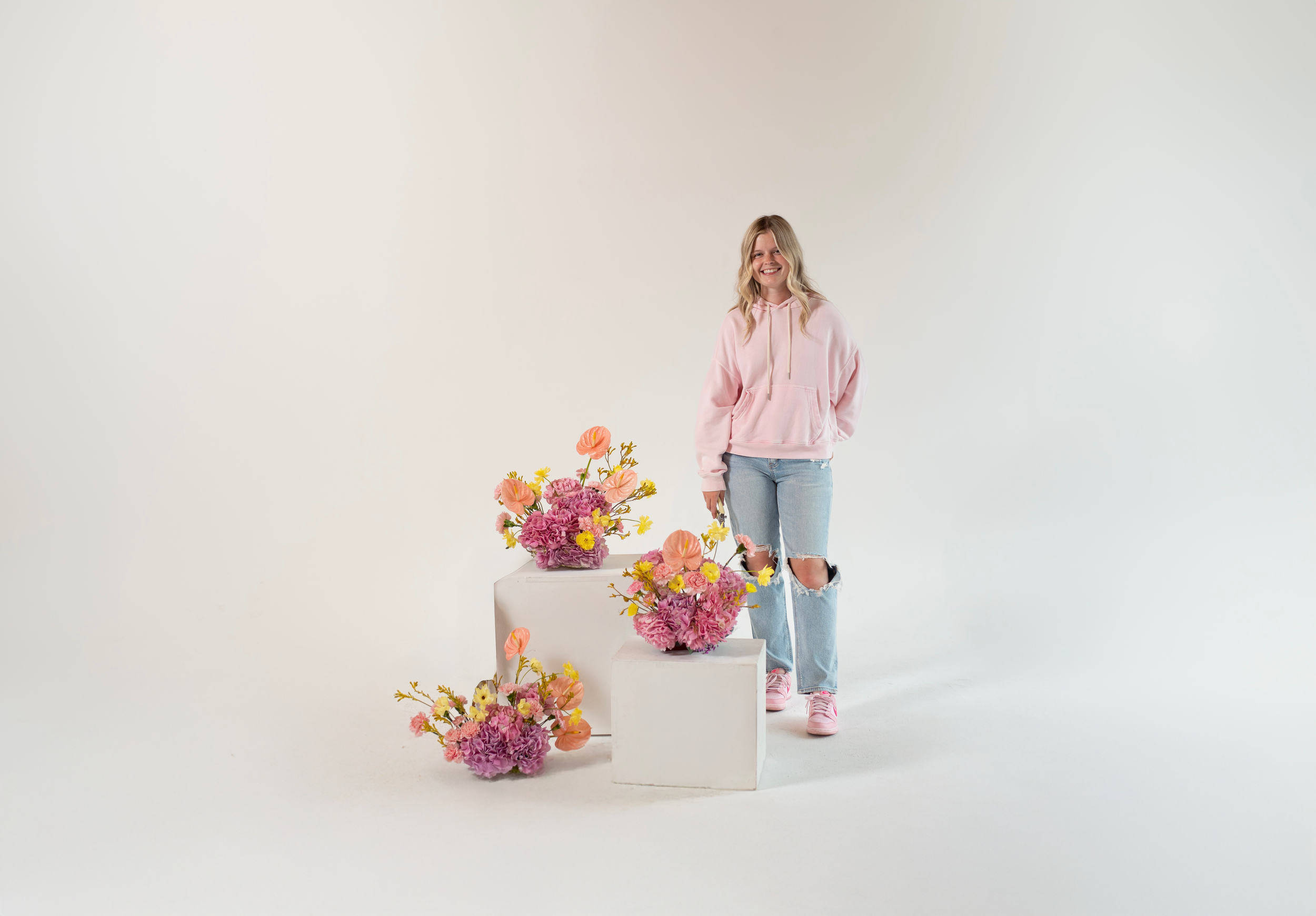 A young woman in a pink hoodie and ripped jeans standing next to white pedestals with colorful pink and yellow flower arrangements on a white background.