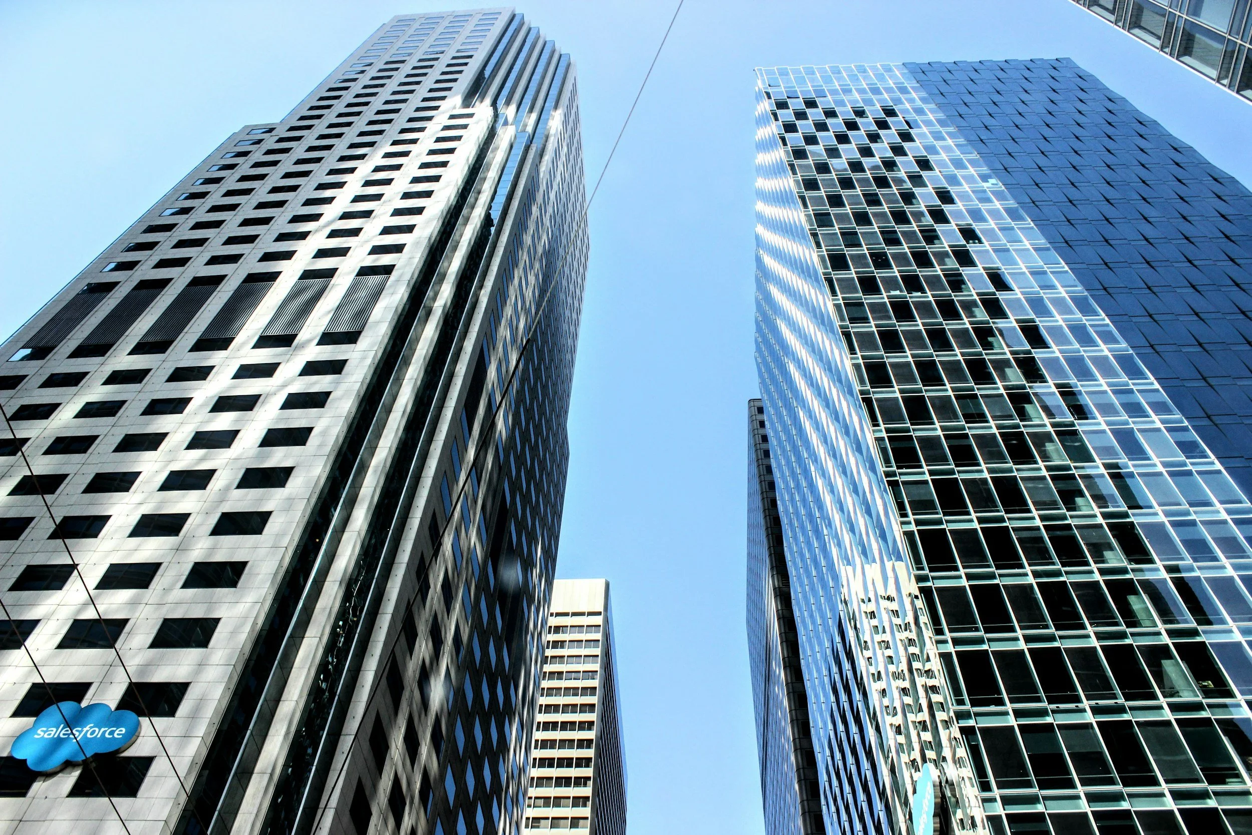 A view of modern glass skyscrapers from below against a clear blue sky, with a Salesforce logo visible on one building.