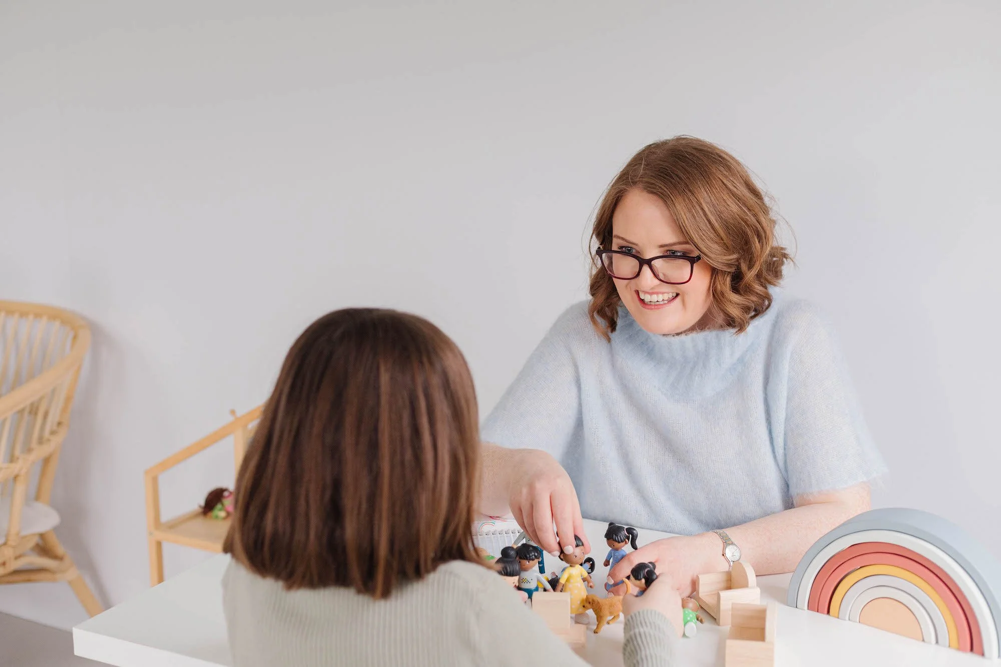 A woman (Jill) with glasses and curly brown hair smiling while playing with a young child with brown hair at a white table. The table has toy figurines, a rainbow-shaped toy, and small wooden blocks.