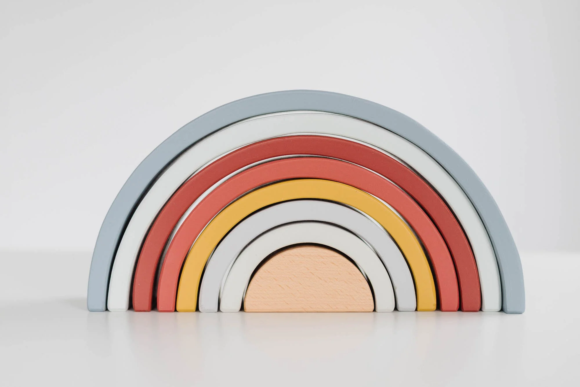 Colourful wooden rainbow toy with five arches in shades of blue, white, red, yellow, and peach, set against a plain white background.