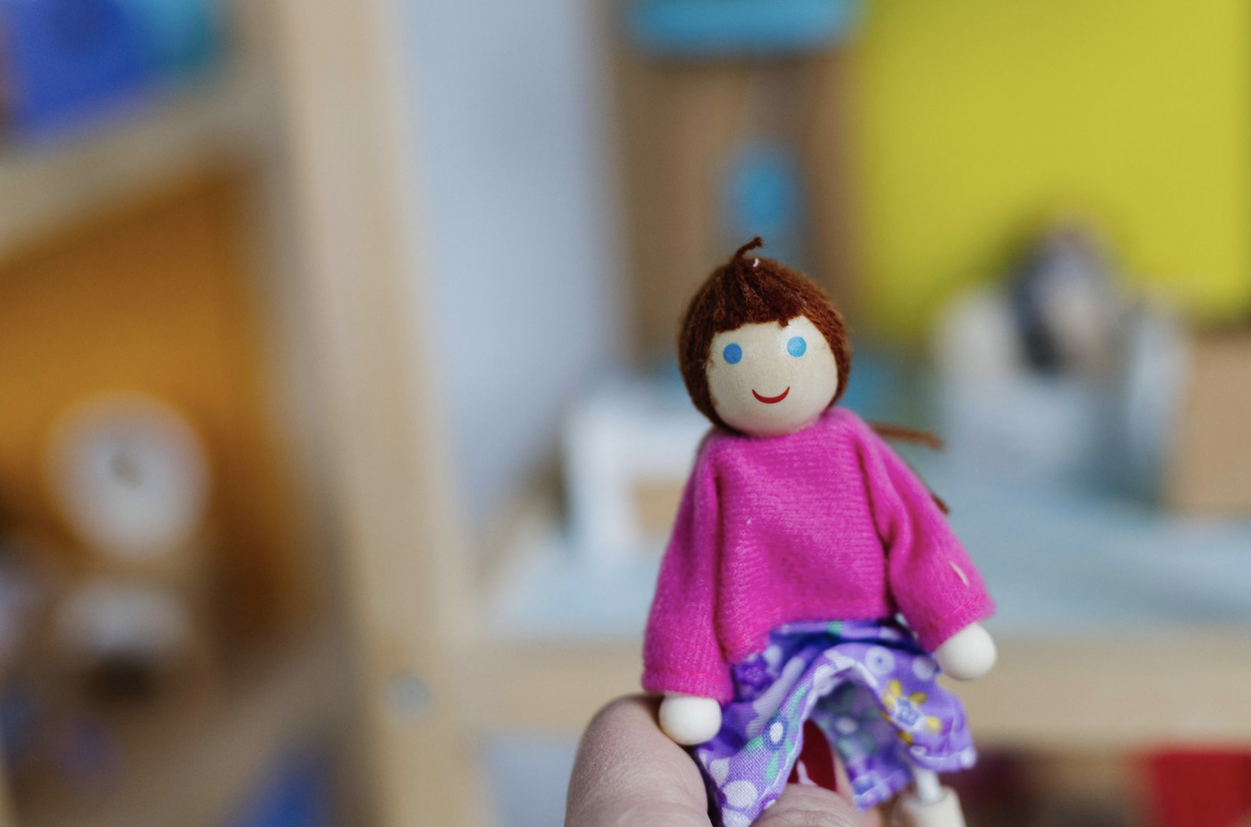A small handmade doll with brown yarn hair, a pink sweater, and purple pants with floral patterns, being held in a person's hand.