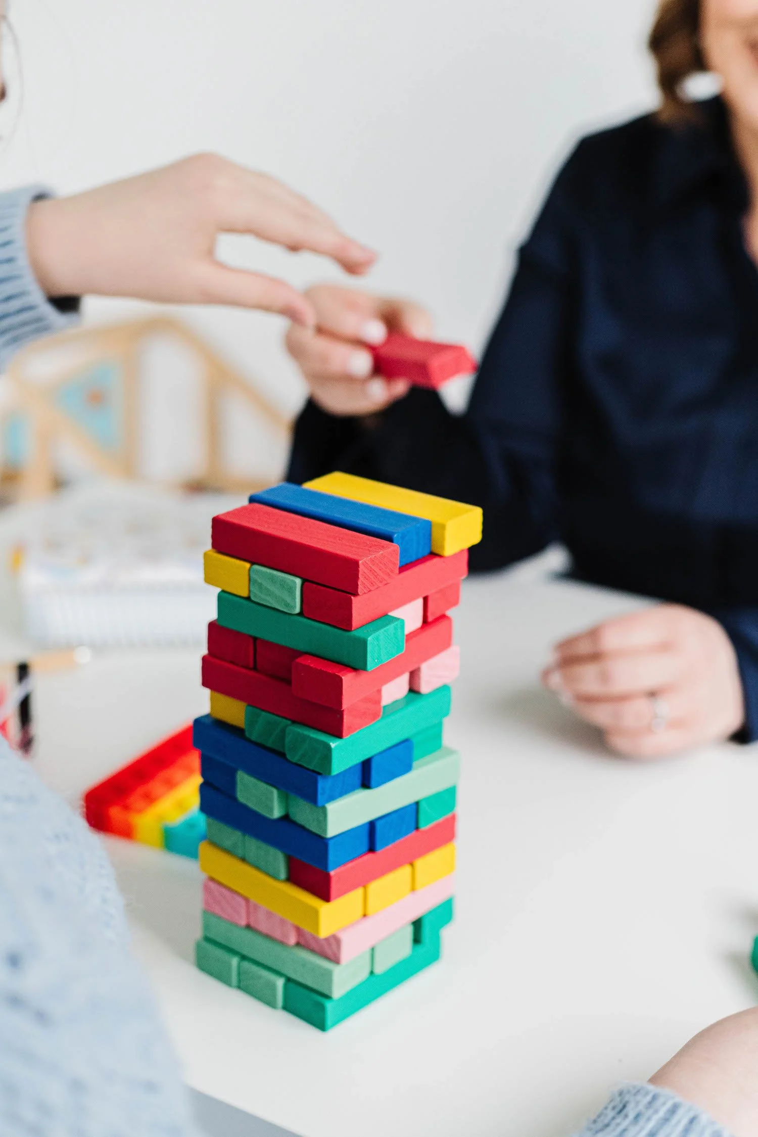 Close-up of a colourful Jenga tower on a white table, with people playing and arranging the blocks.