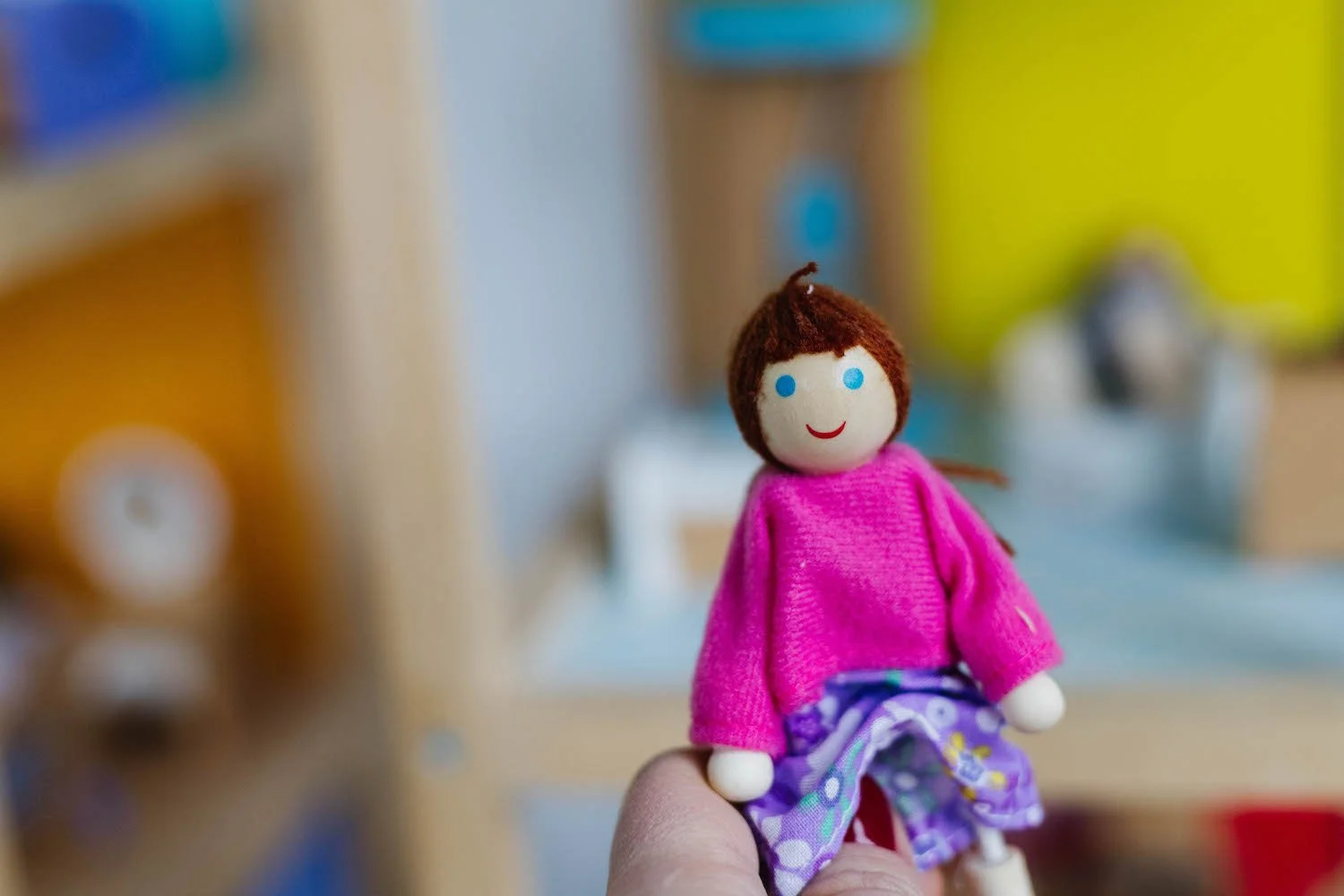 A small doll with a smiling face, brown hair, wearing a pink top and purple patterned pants, held by a person's hand. The background is blurred with colourful objects.