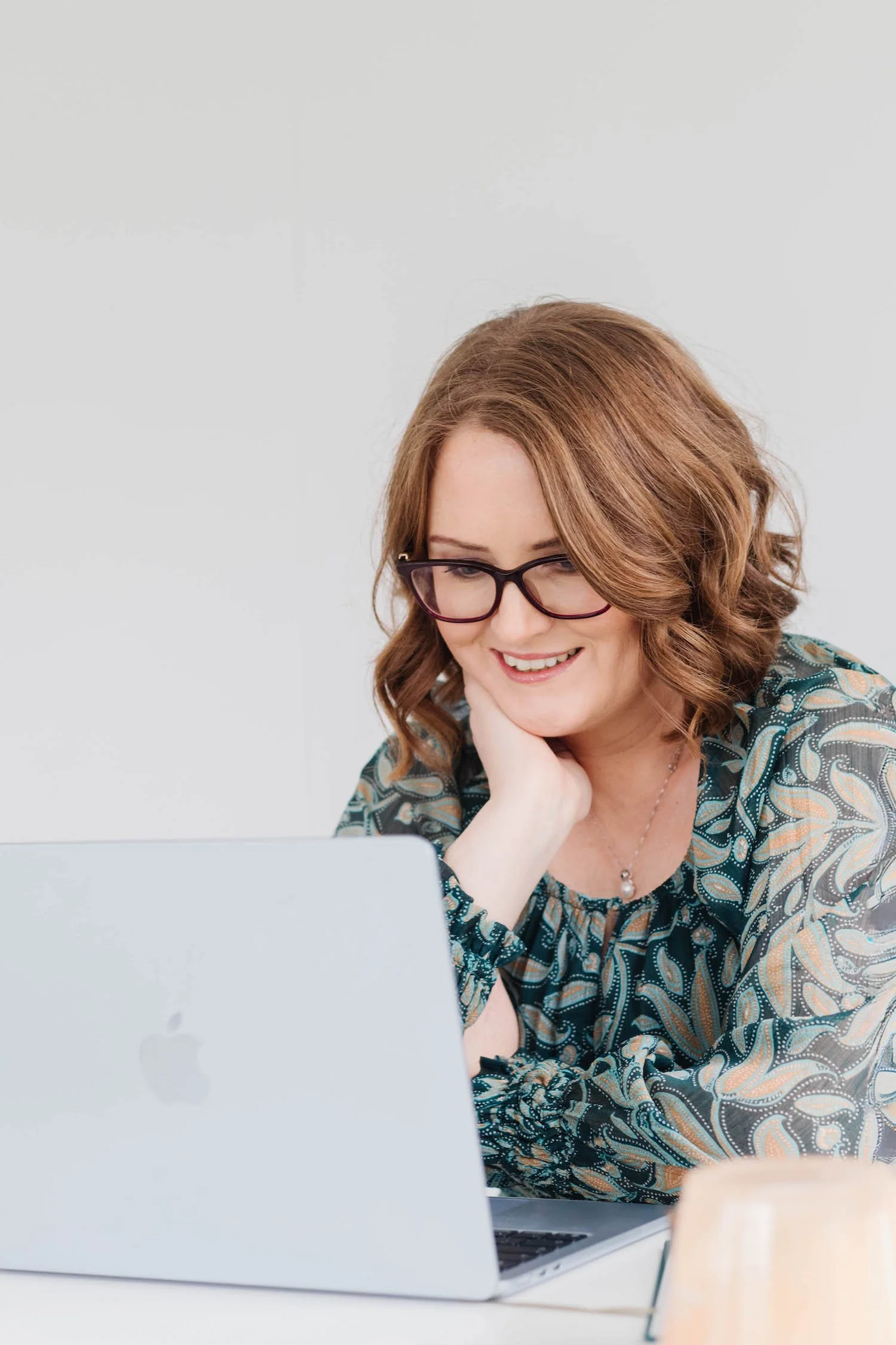A woman with red hair and glasses smiling while looking at a laptop.