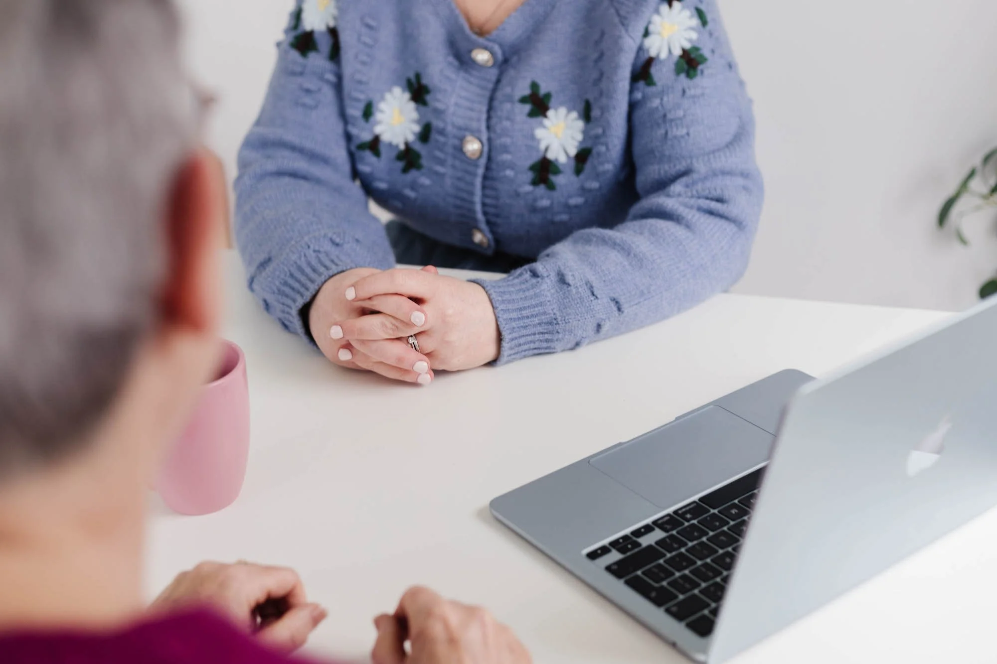 Two people sitting at a white table, one in a purple top with their hair visible, the other in a blue sweater with daisy embroidery, hands clasped, with a silver MacBook on the table. A pink mug is also on the table.