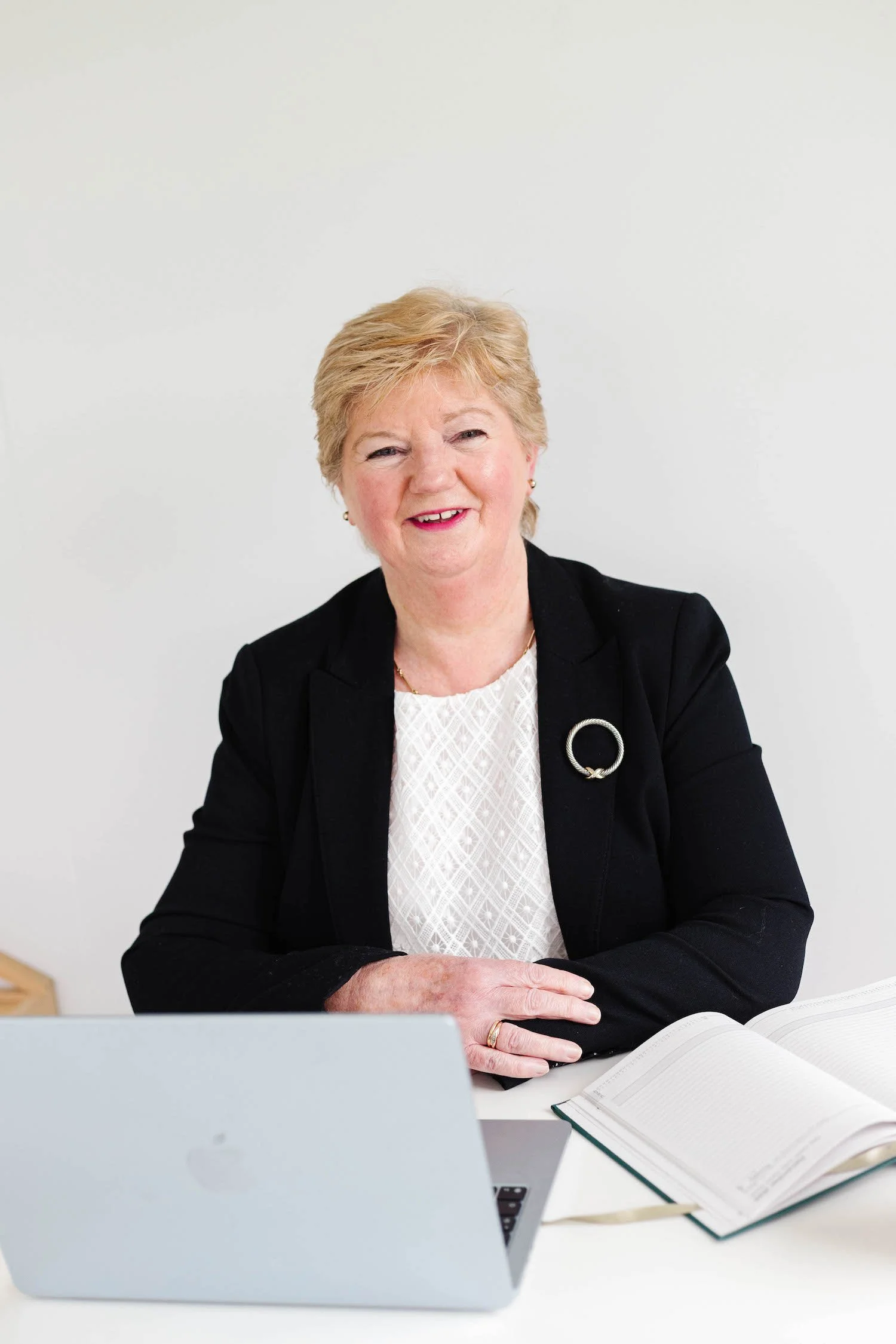 A smiling woman with short blonde hair sitting at a desk, with a silver laptop and an open notebook in front of her, against a plain white background.
