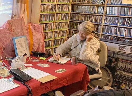 A volunteer sits at a desk in a room filled with shelves of CDs, writing in a notebook with a phone held to her ear during public radio pledge drive.