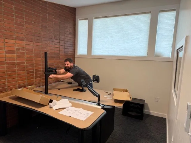 A man setting up a computer monitor on a desk in an office with a brick wall and large windows.