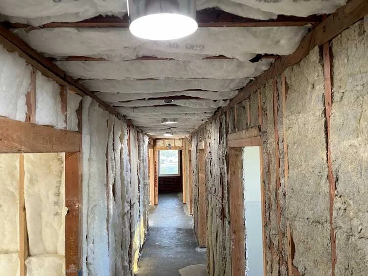 Construction hallway with insulation in unfinished walls, exposed wooden framing, and a ceiling with insulation. A door frame at the end leads to a room with a window, and a ceiling light fixture is visible.