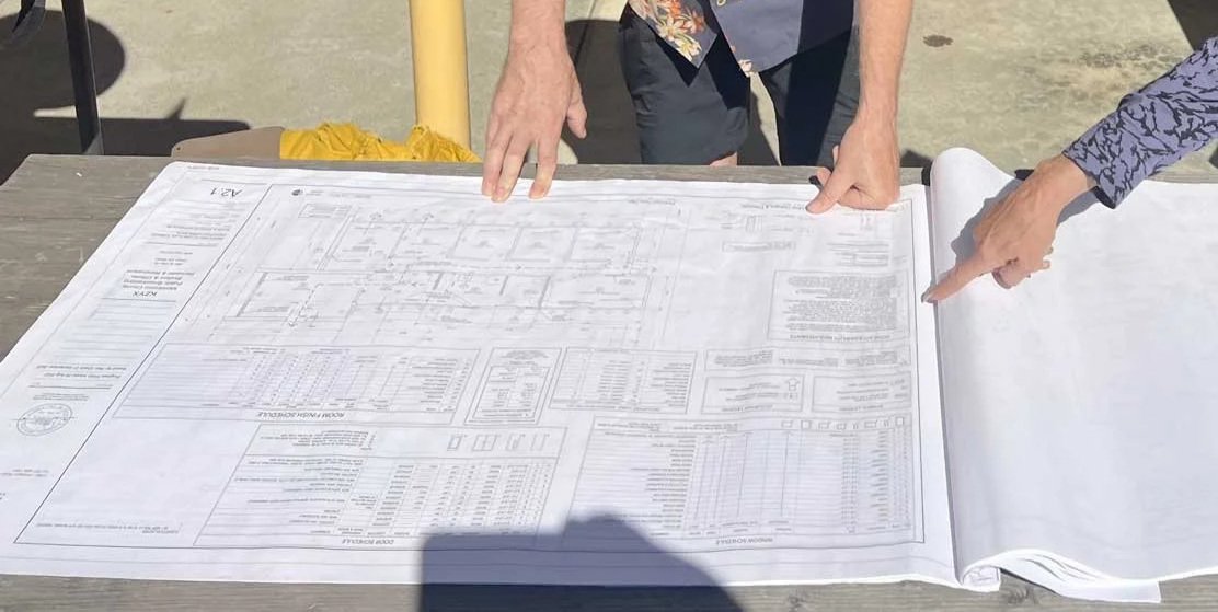 Two people examining and pointing at architectural blueprints spread out on a wooden table outdoors.