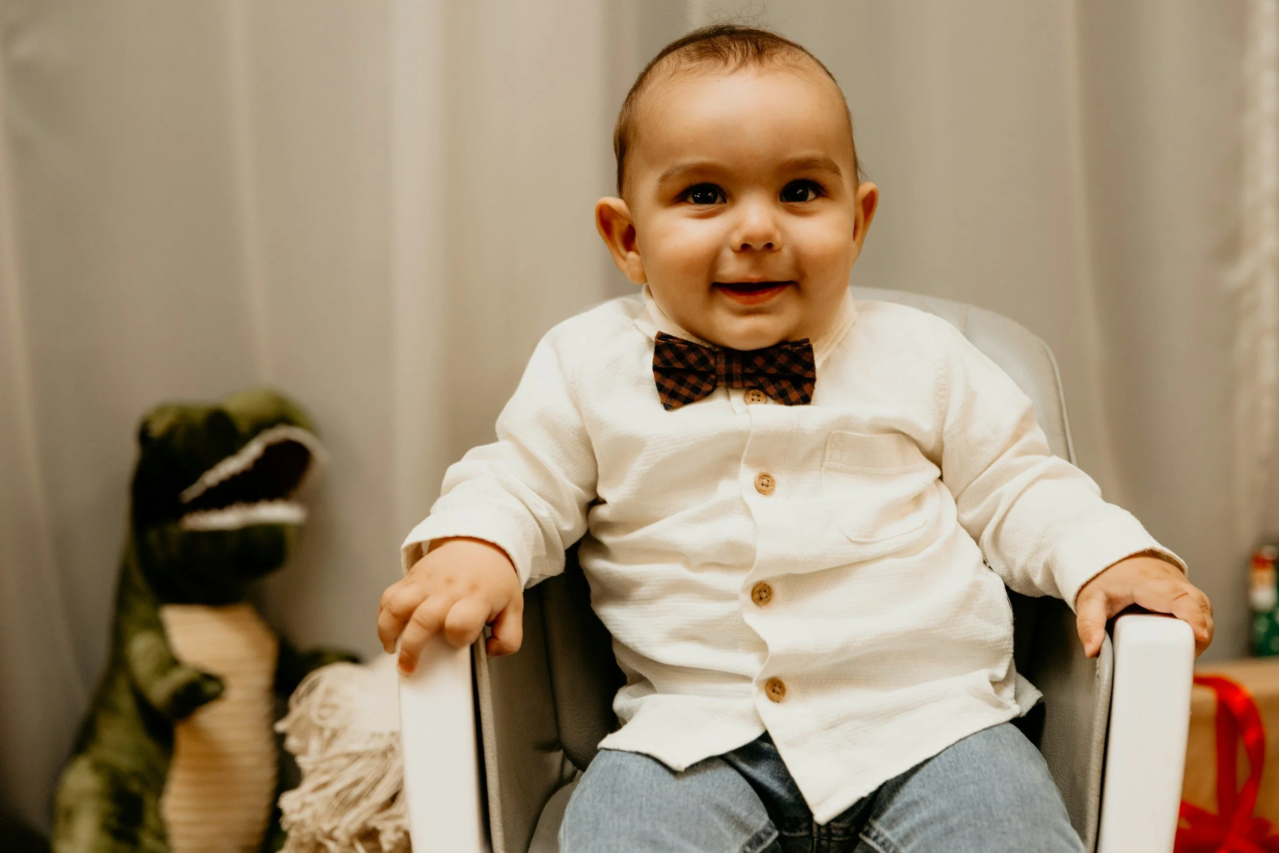 A smiling young boy sitting in a white plastic chair with a dinosaur toy in the background.