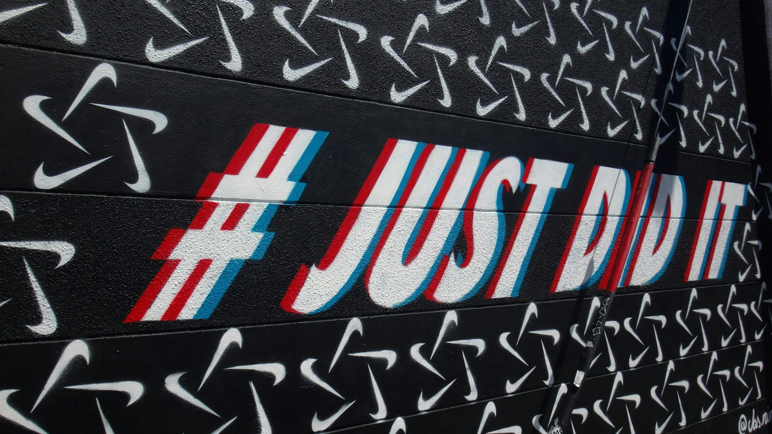 Street art mural featuring the Nike logo pattern in white and the hashtag #JUSTDOIT in red, white, and blue with a bicycle handlebar in front.