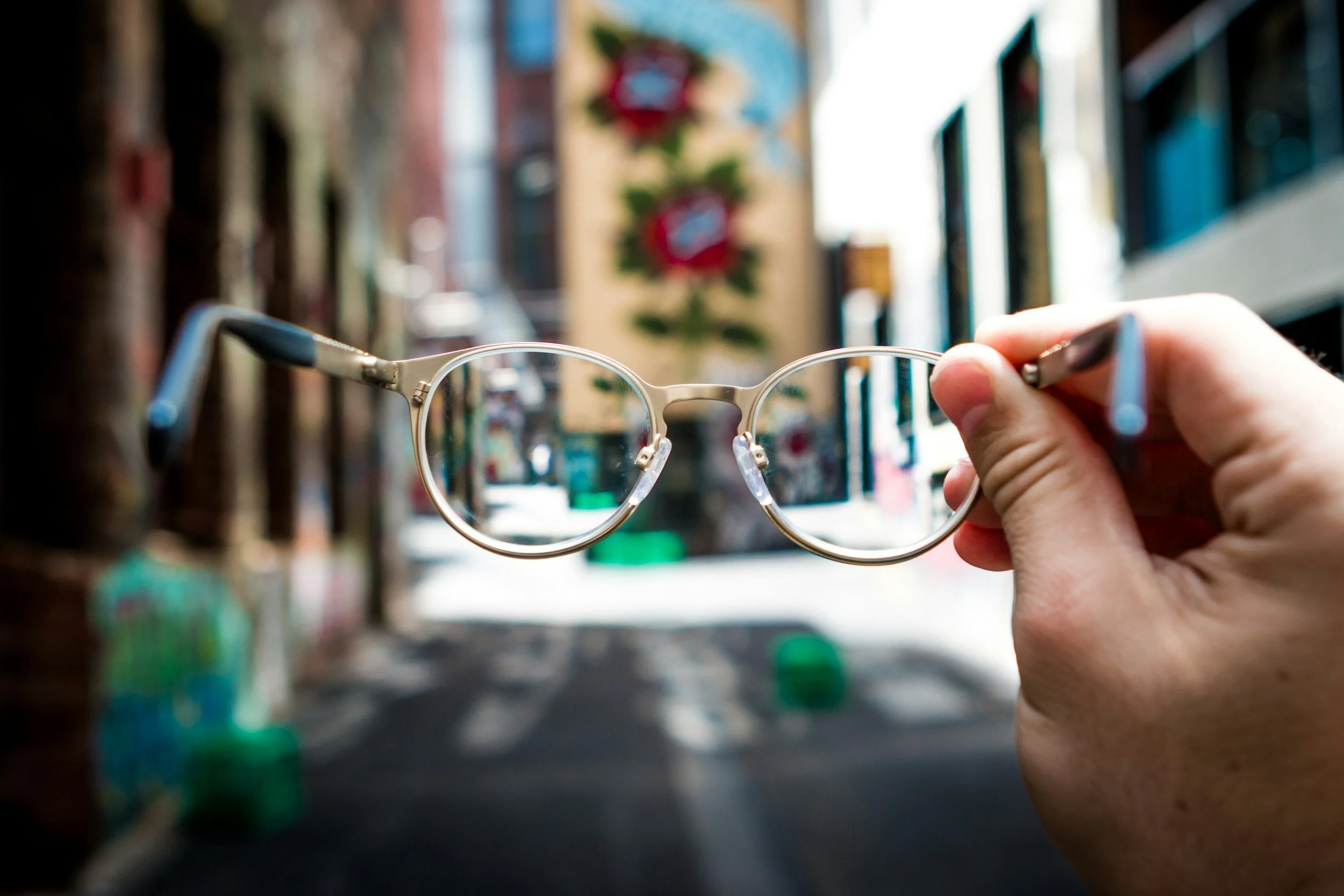 Hand holding glasses in front of an urban street scene with buildings and a mural.
