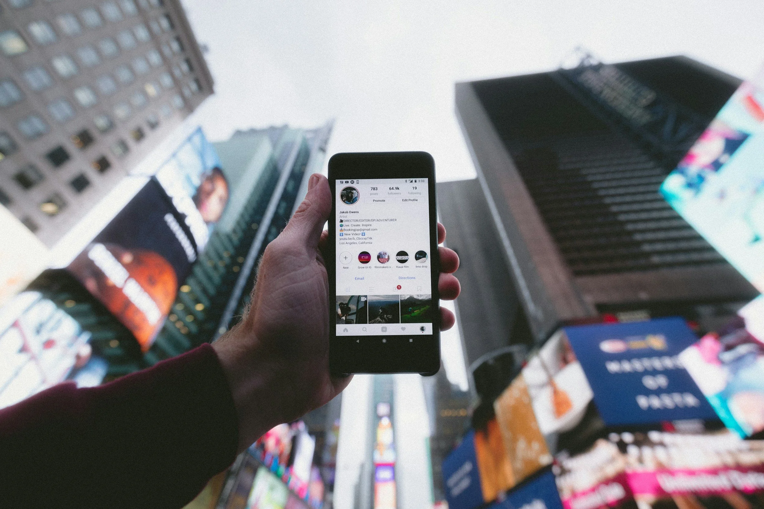 Person holding a smartphone with an Instagram profile open, surrounded by tall buildings and digital billboards in Times Square, New York City.