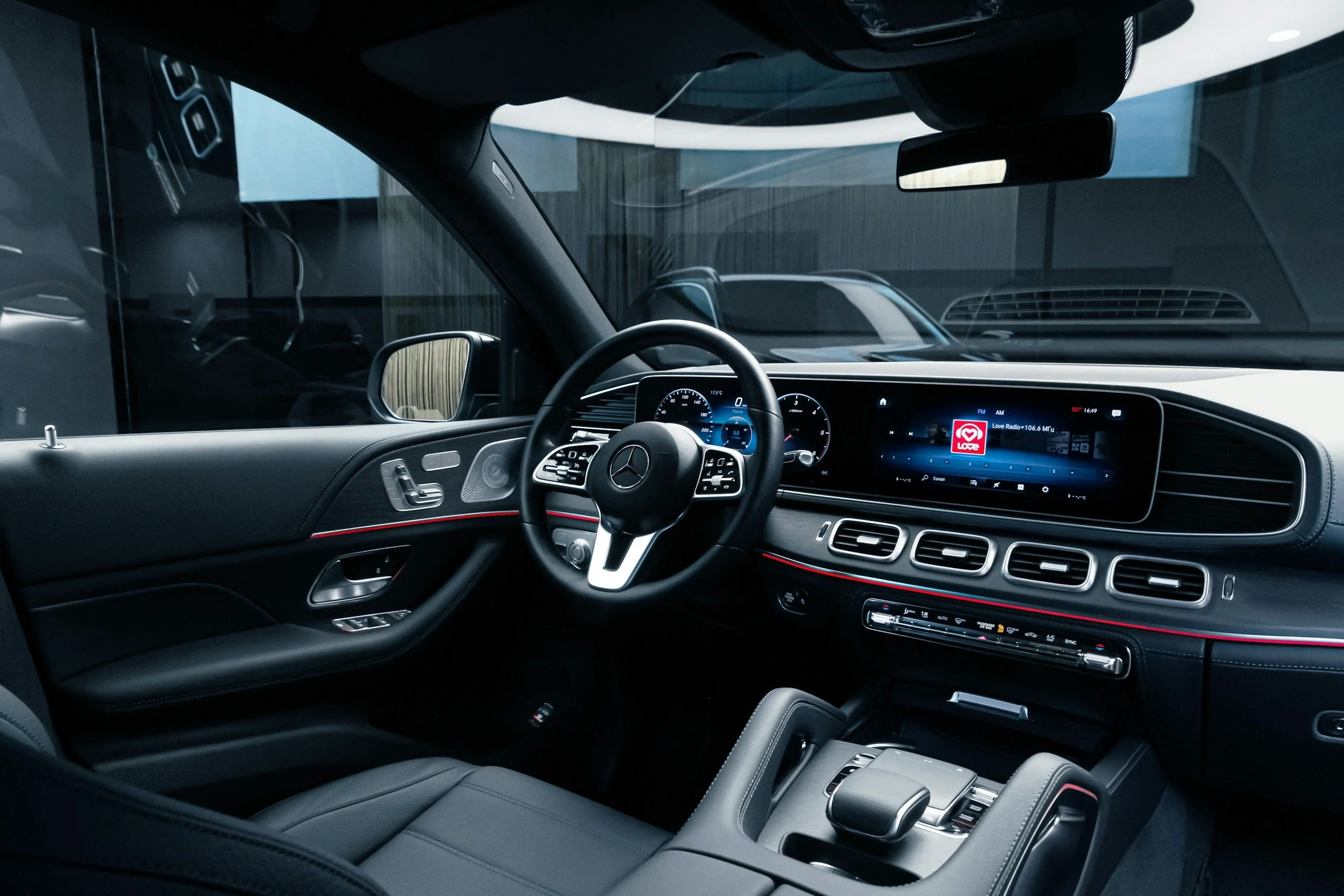 Interior of a modern Mercedes-Benz vehicle, featuring a digital dashboard, steering wheel with controls, a central infotainment screen displaying a radio station, and black leather seats.
