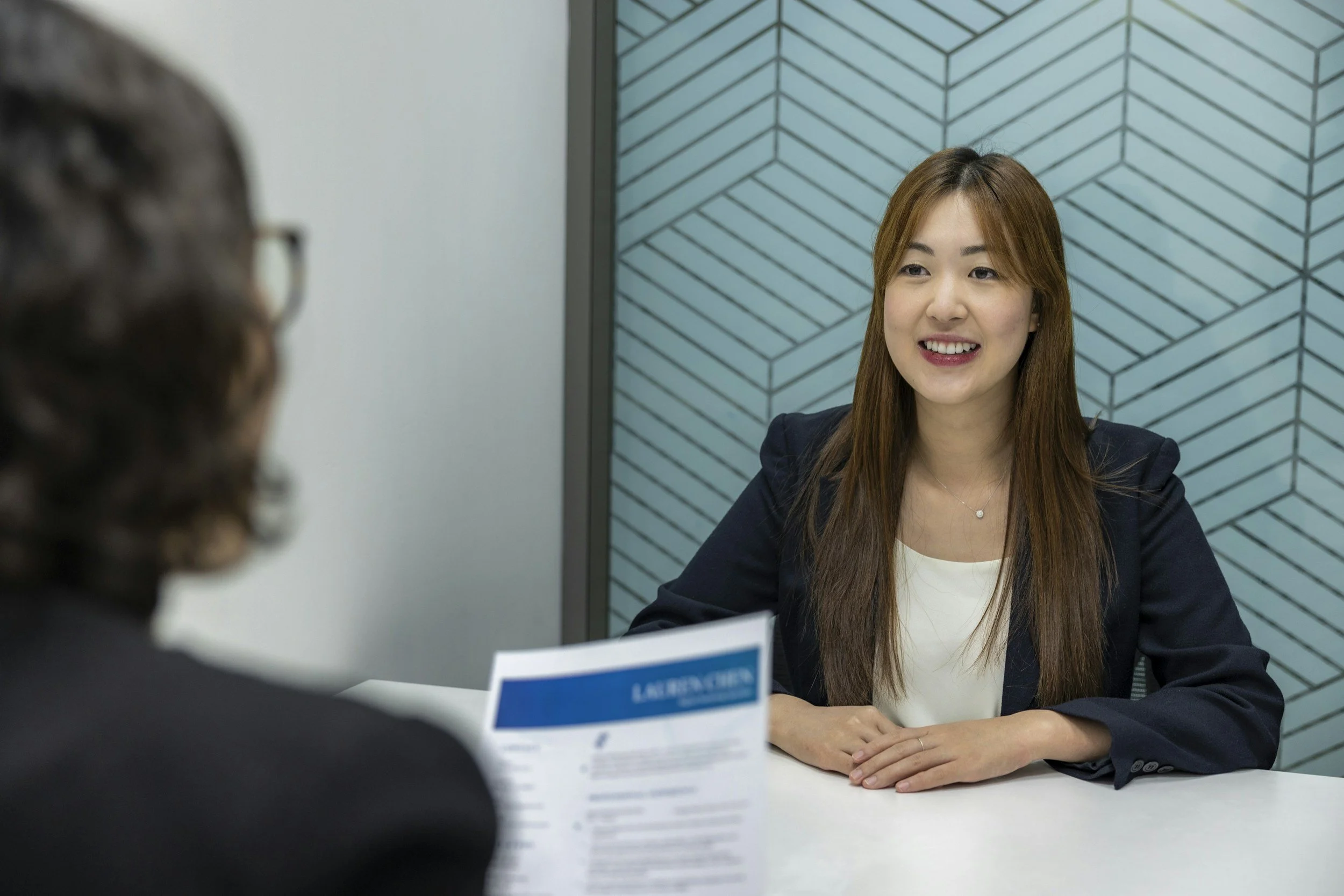 A woman with long brown hair, wearing a navy blazer and white top, sitting at a table during a job interview or meeting, smiling and engaging in conversation with a person whose back is to the camera holding a document.