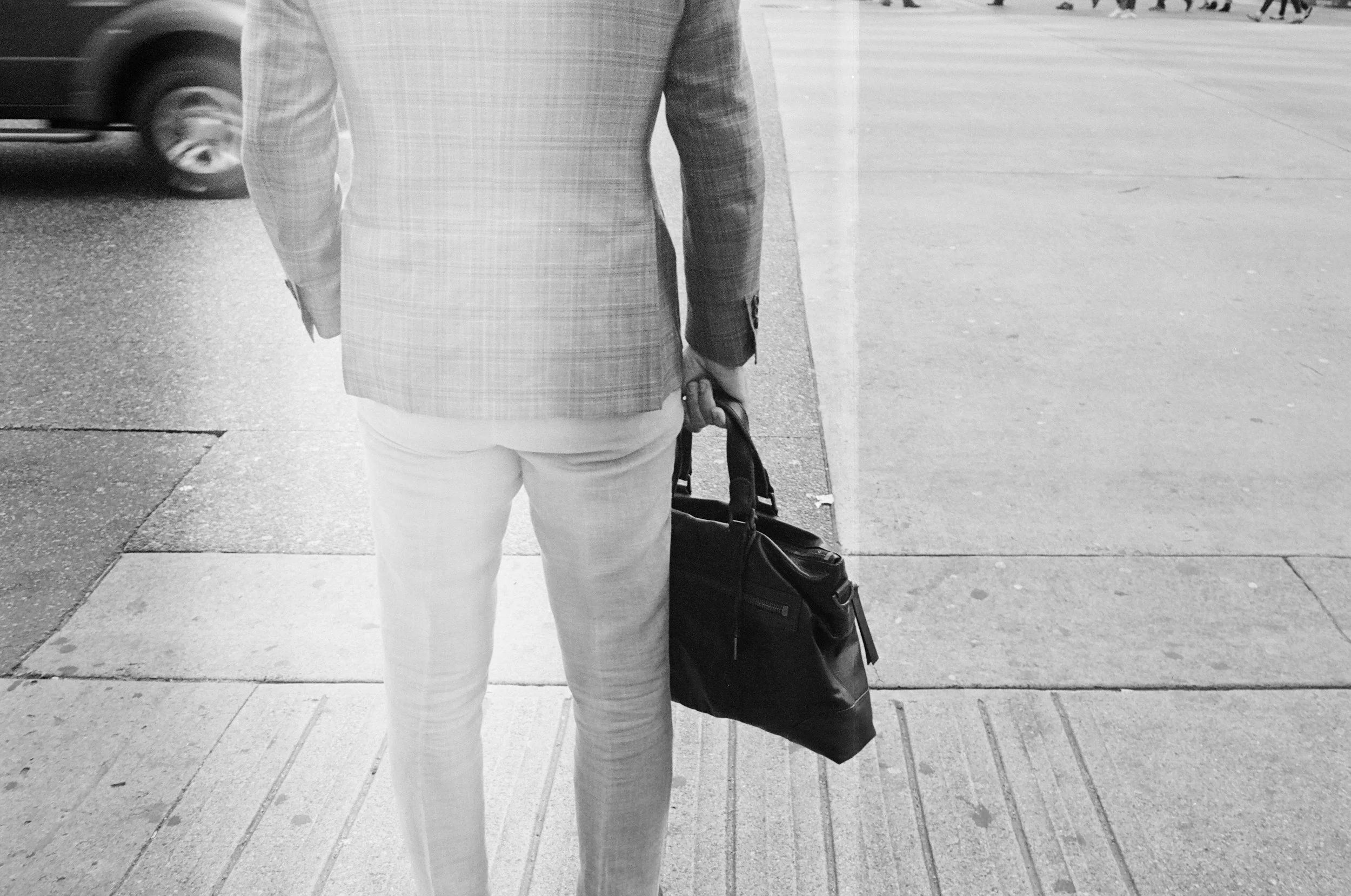 Back view of a man in light-colored pants and a checked blazer holding a black bag, standing on a sidewalk in an urban area.
