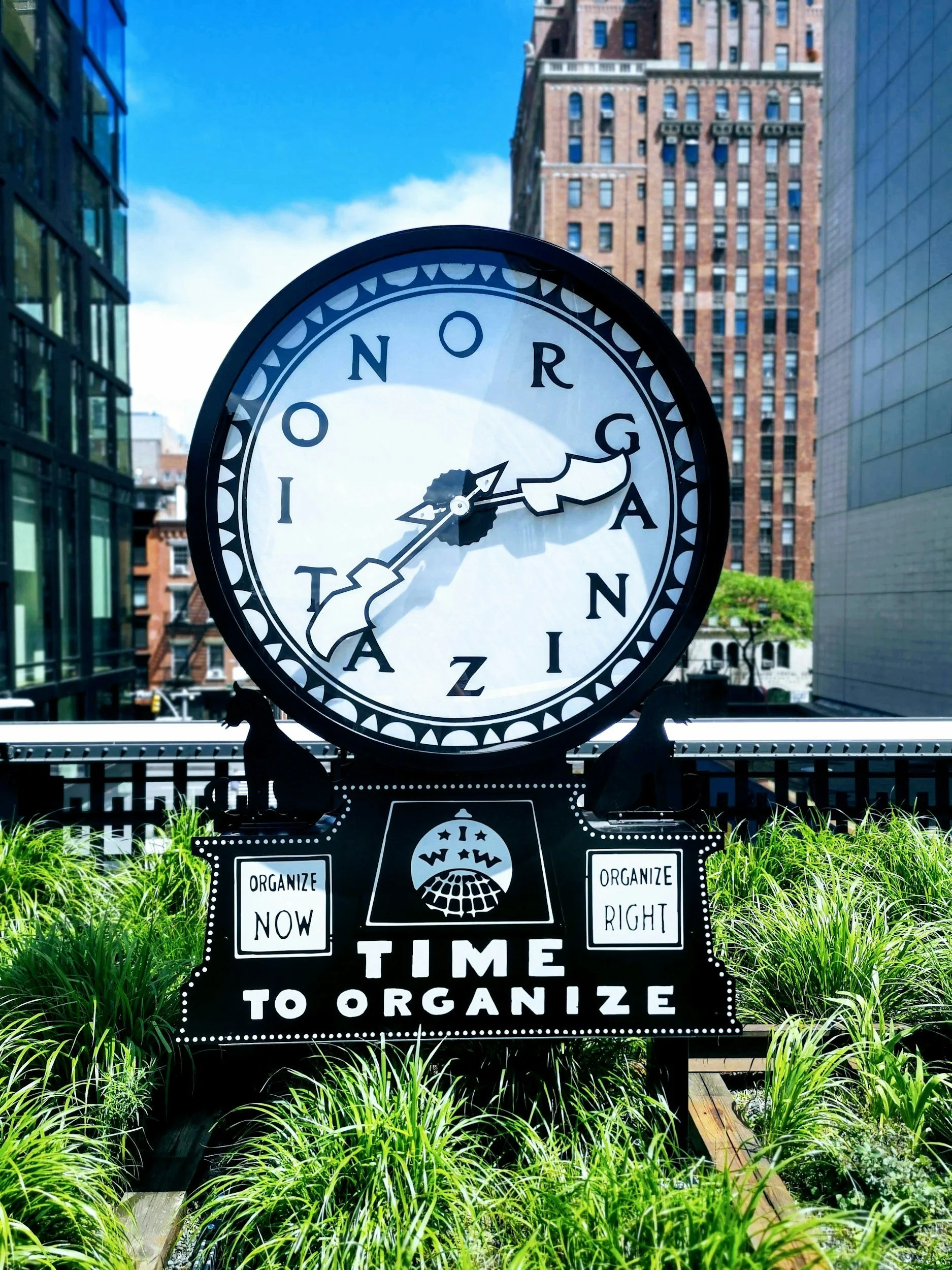 A large round clock with the words 'Ontario, Canada' on its face, showing the time as approximately 3:15. The clock is outdoors, surrounded by green plants, with tall buildings in the background. There is a black and white sign below the clock that reads 'Time to Organize' and has additional smaller signs saying 'Organize Now' and 'Organize Right'.