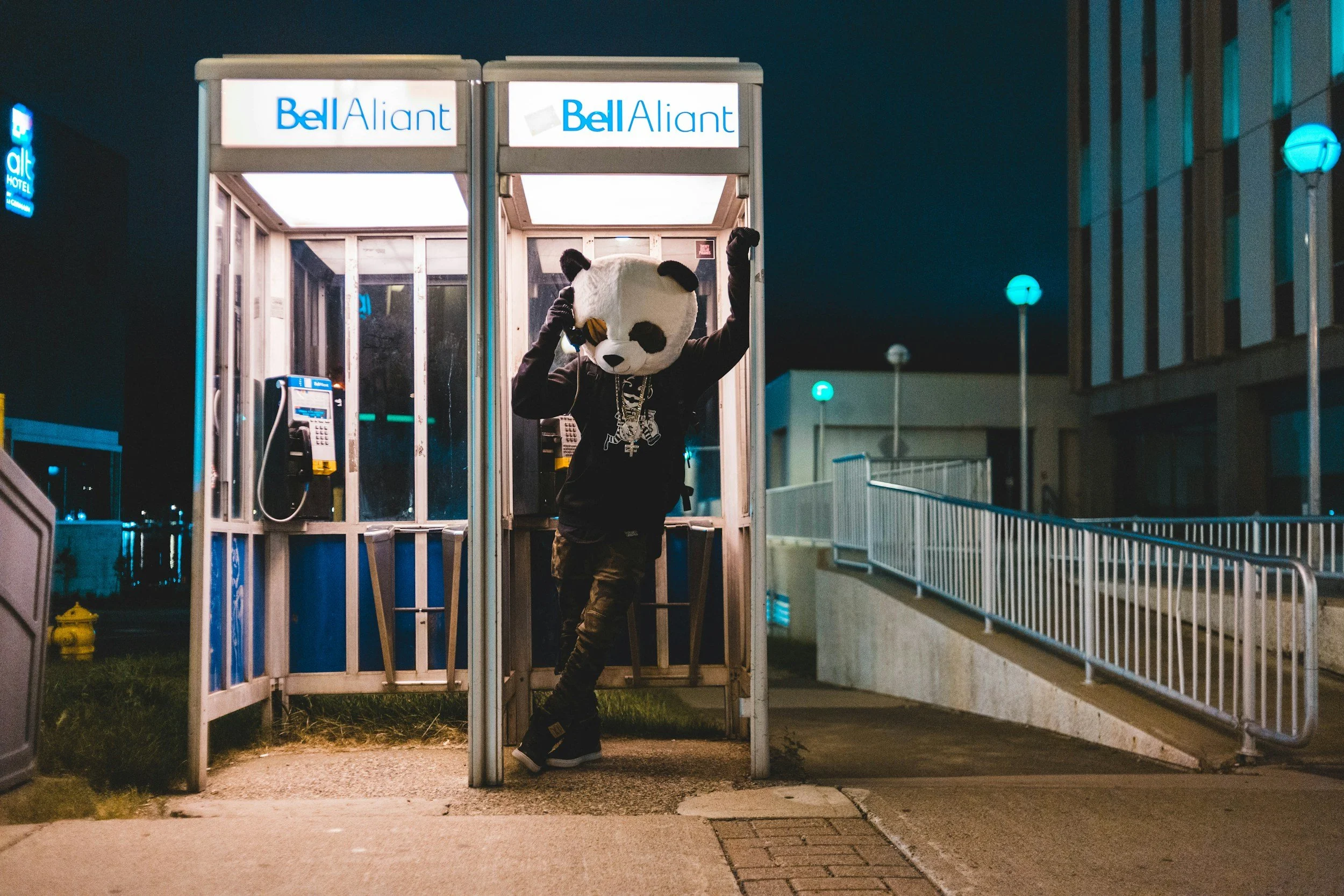Person wearing a panda mask, black hoodie with chains, standing outside a phone booth at night, raising one arm.