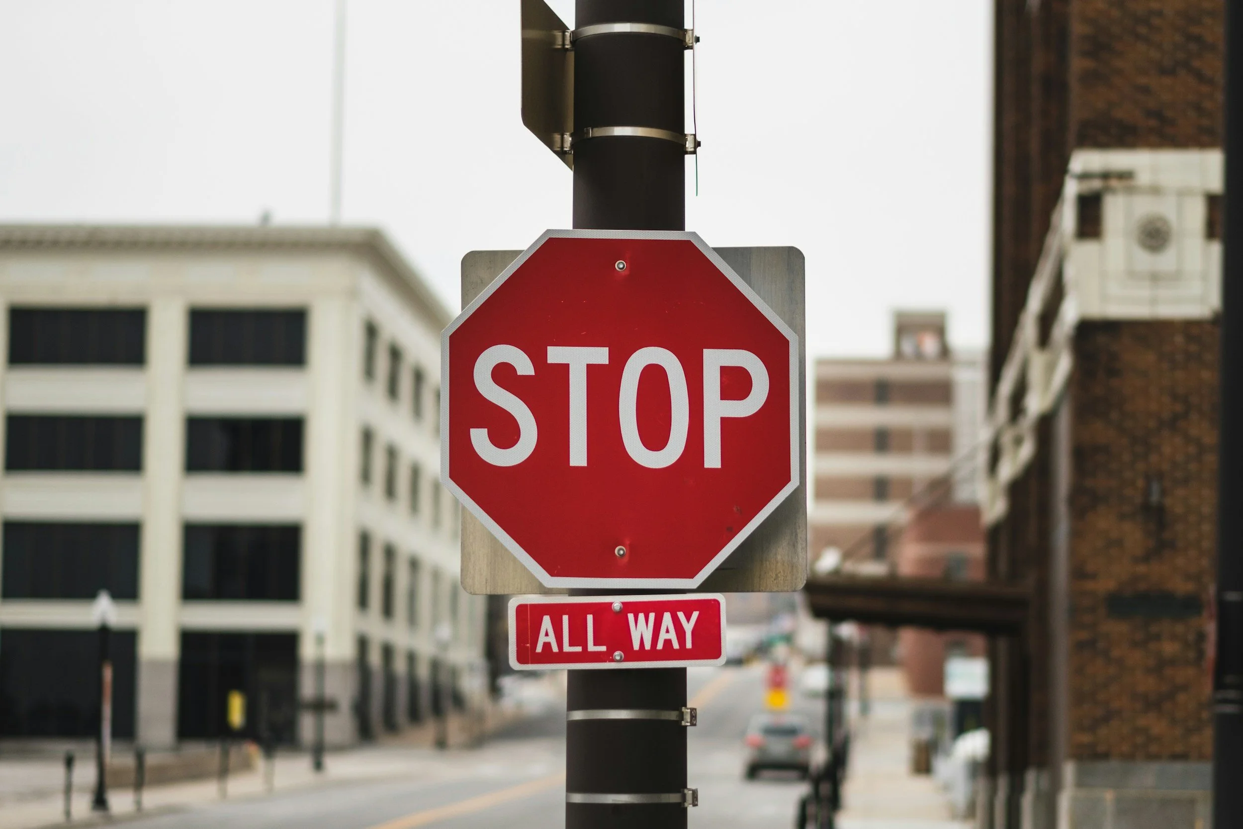 A red stop sign with white letters on a pole in an urban setting with buildings and a street in background.