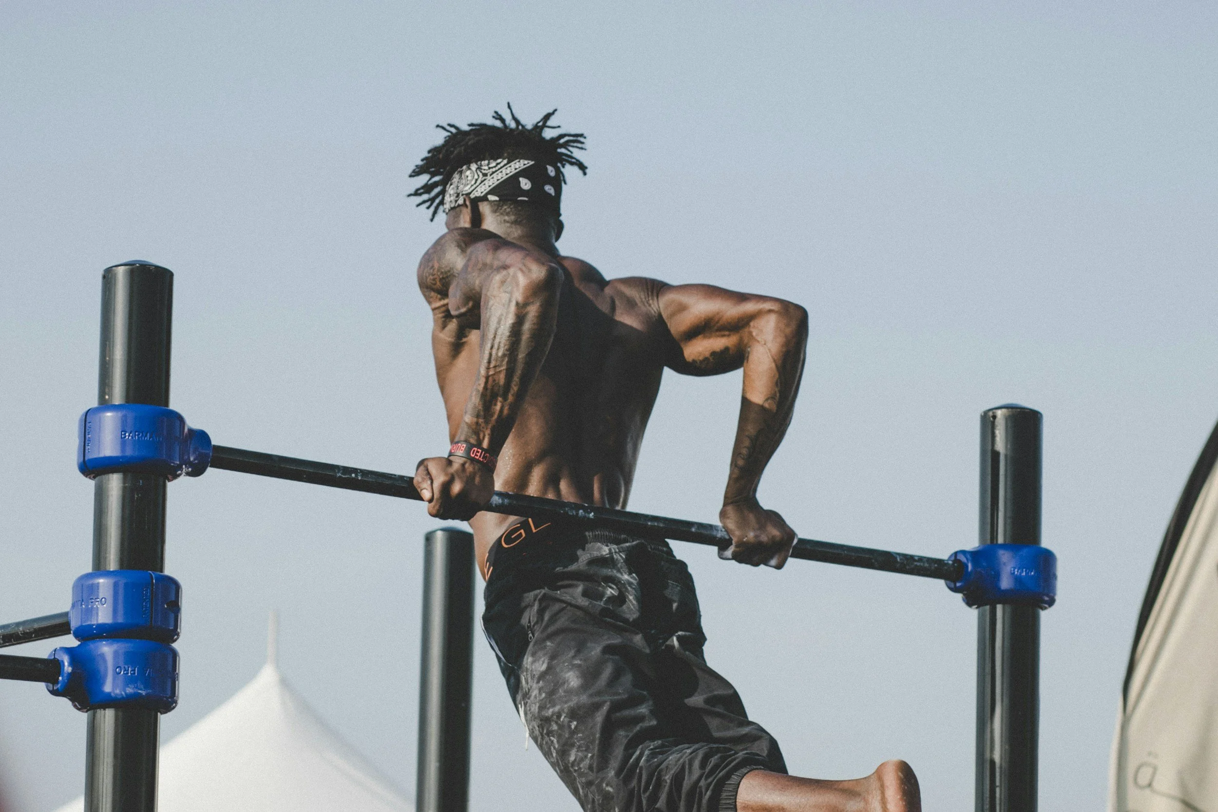 A shirtless man with tattoos performing a calisthenics exercise on outdoor bars, wearing a bandana and black shorts.