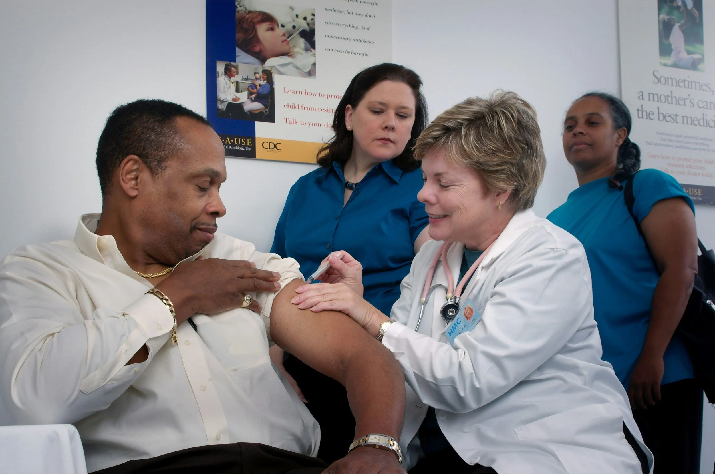 A woman receives a vaccination shot from a healthcare professional while sitting in a chair, with two women standing nearby observing, in a clinic setting with health posters on the wall.