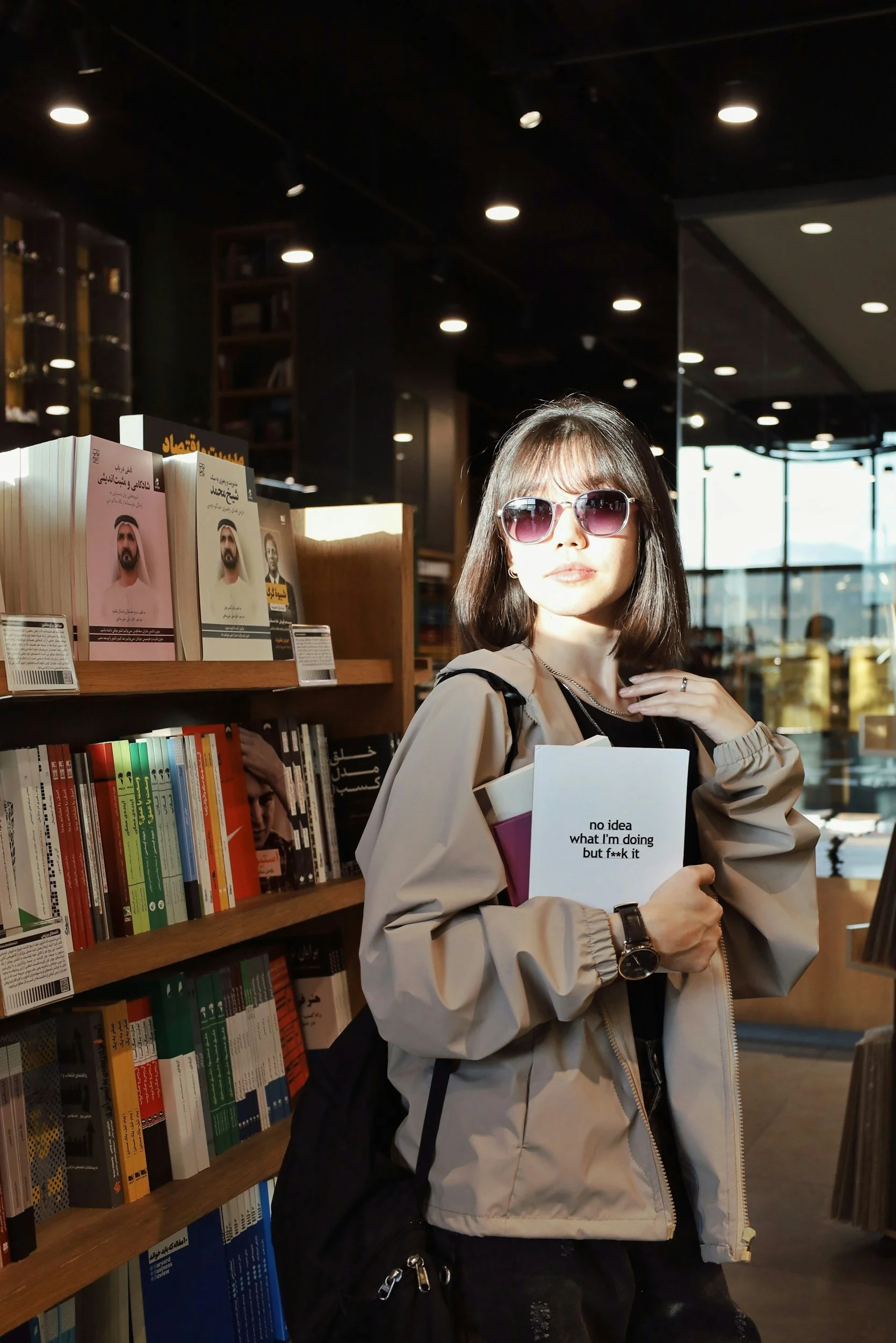 A young woman with shoulder-length dark hair wearing sunglasses, a beige jacket, and a watch stands in a bookstore holding a book titled "no idea what I'm doing but f**k it." She is looking off to the side with sunlight illuminating her face. Behind her are shelves filled with books, and large windows allow natural light to enter.
