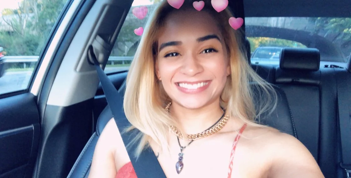 A smiling woman with blonde hair wearing a red top, sitting in a car with hearts overlay filter.