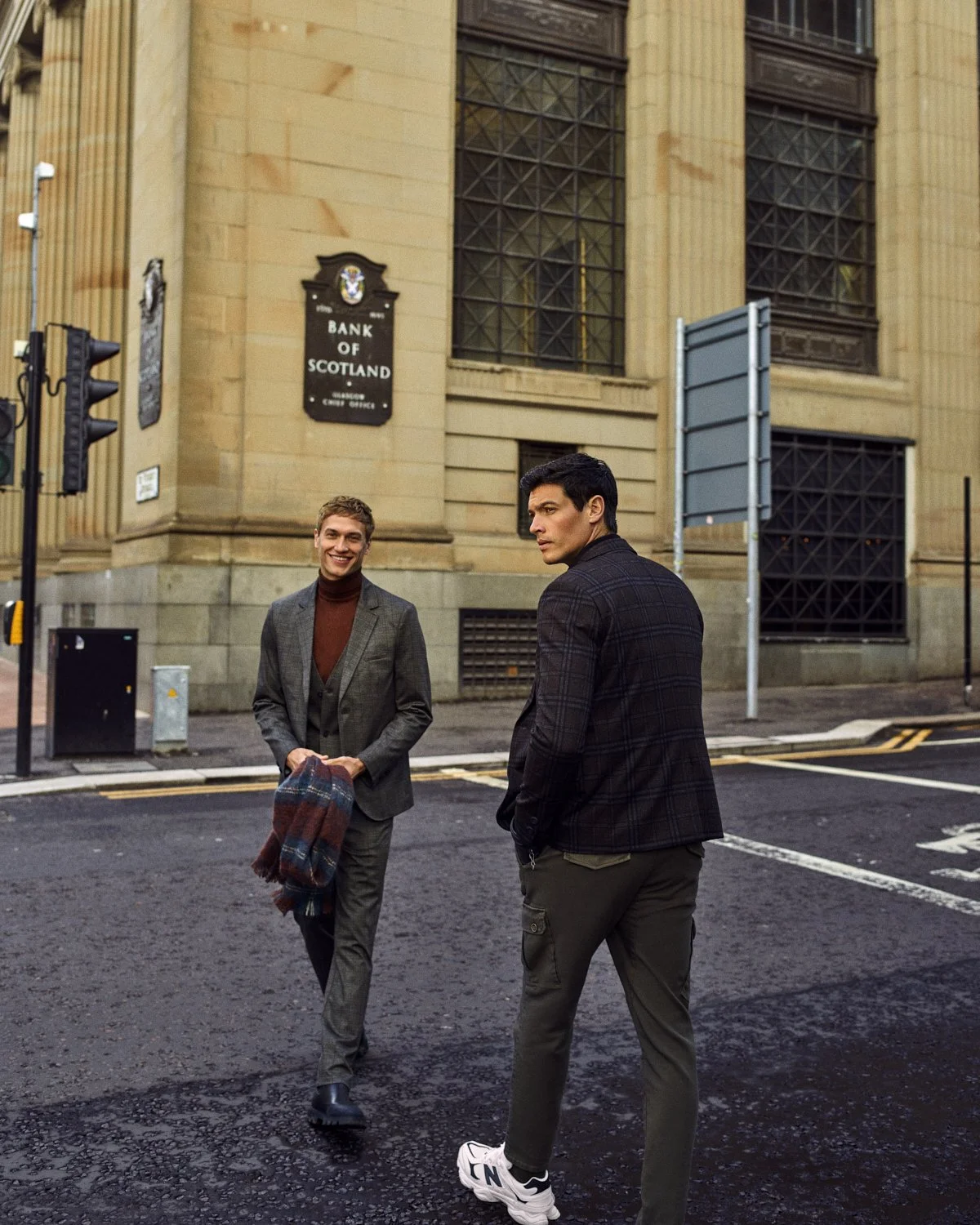 Two men walking on a city street in front of the Bank of Scotland building, one smiling and holding a plaid scarf, the other looking away, both dressed in stylish jackets.