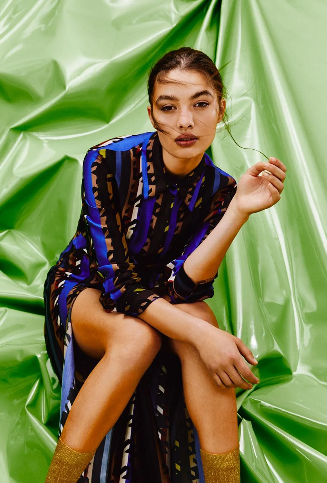 A young woman sitting with crossed legs against a shiny, crinkled green background. She is wearing a colorful, patterned dress with blue, black, and beige tones, and gold socks. She has brown hair, some strands across her face, and is looking at the camera with a neutral expression.