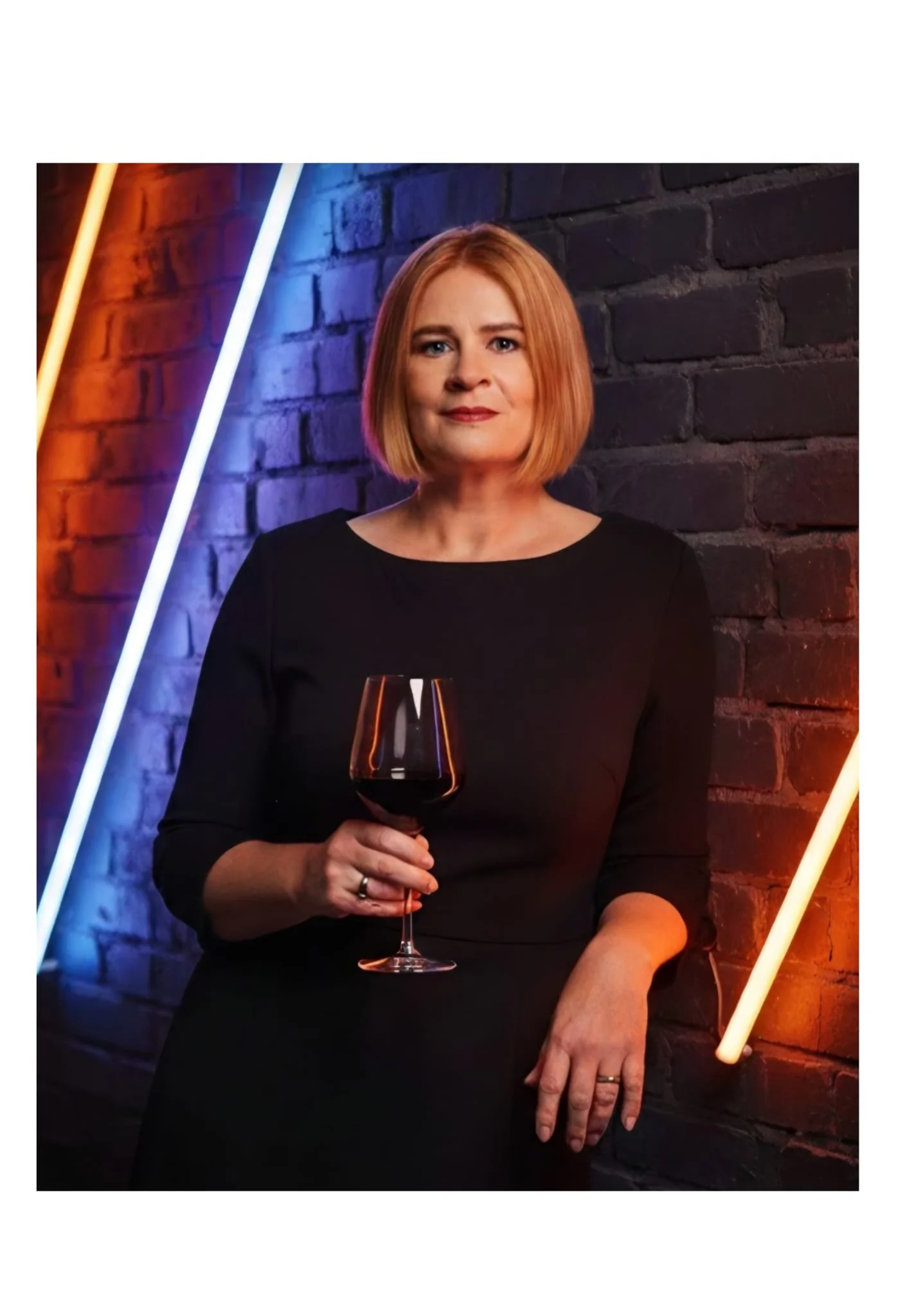 A woman with short red hair, wearing a black dress, holding a glass of red wine, standing in front of a brick wall illuminated with colorful neon lights.