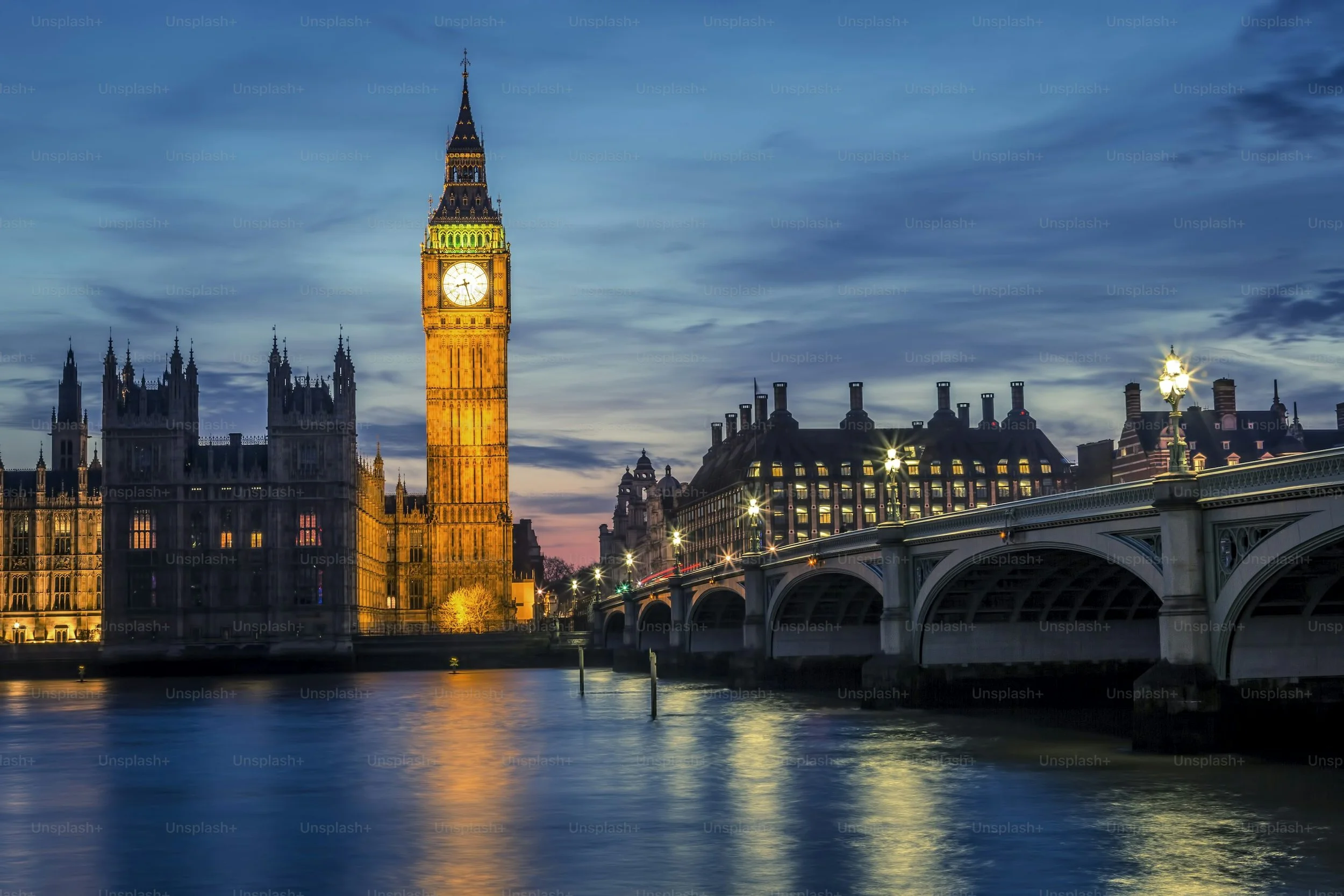 London's Big Ben clock tower illuminated at dusk, with the Houses of Parliament, a bridge with street lamps, and a river reflecting the city lights.