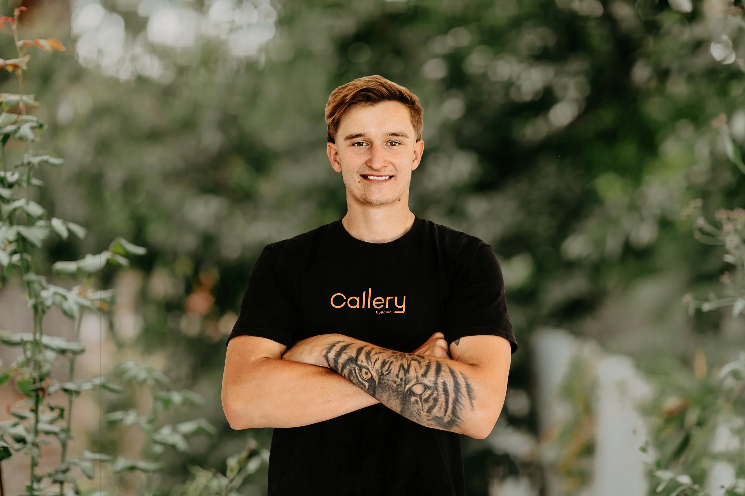 A young man with blond hair and tattoos on his left arm, smiling with arms crossed, wearing a black t-shirt with orange and white text that reads "Callery building".