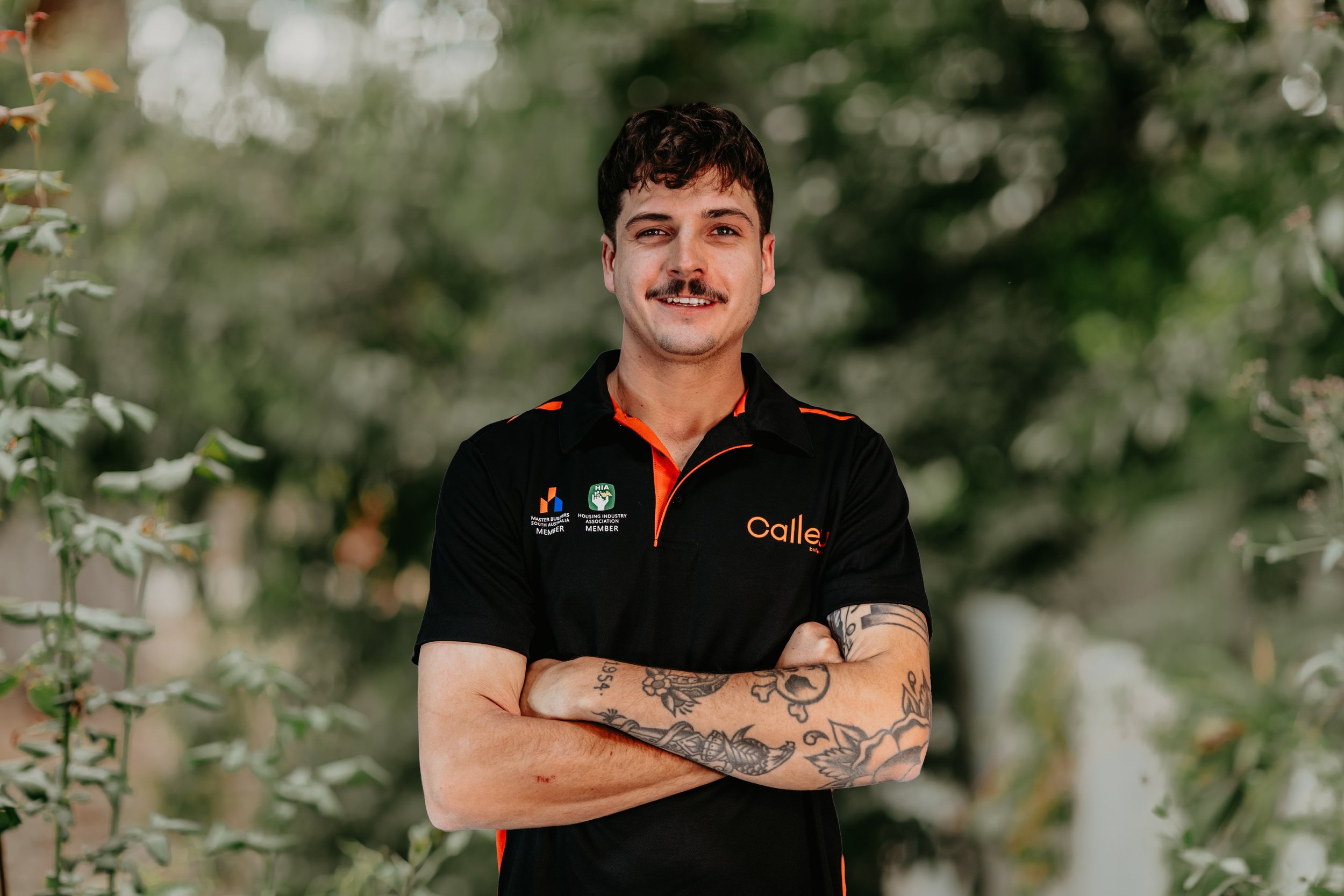 A man wearing a black and orange work shirt and gray gloves stands in a construction site, looking down at a block of stone or concrete.