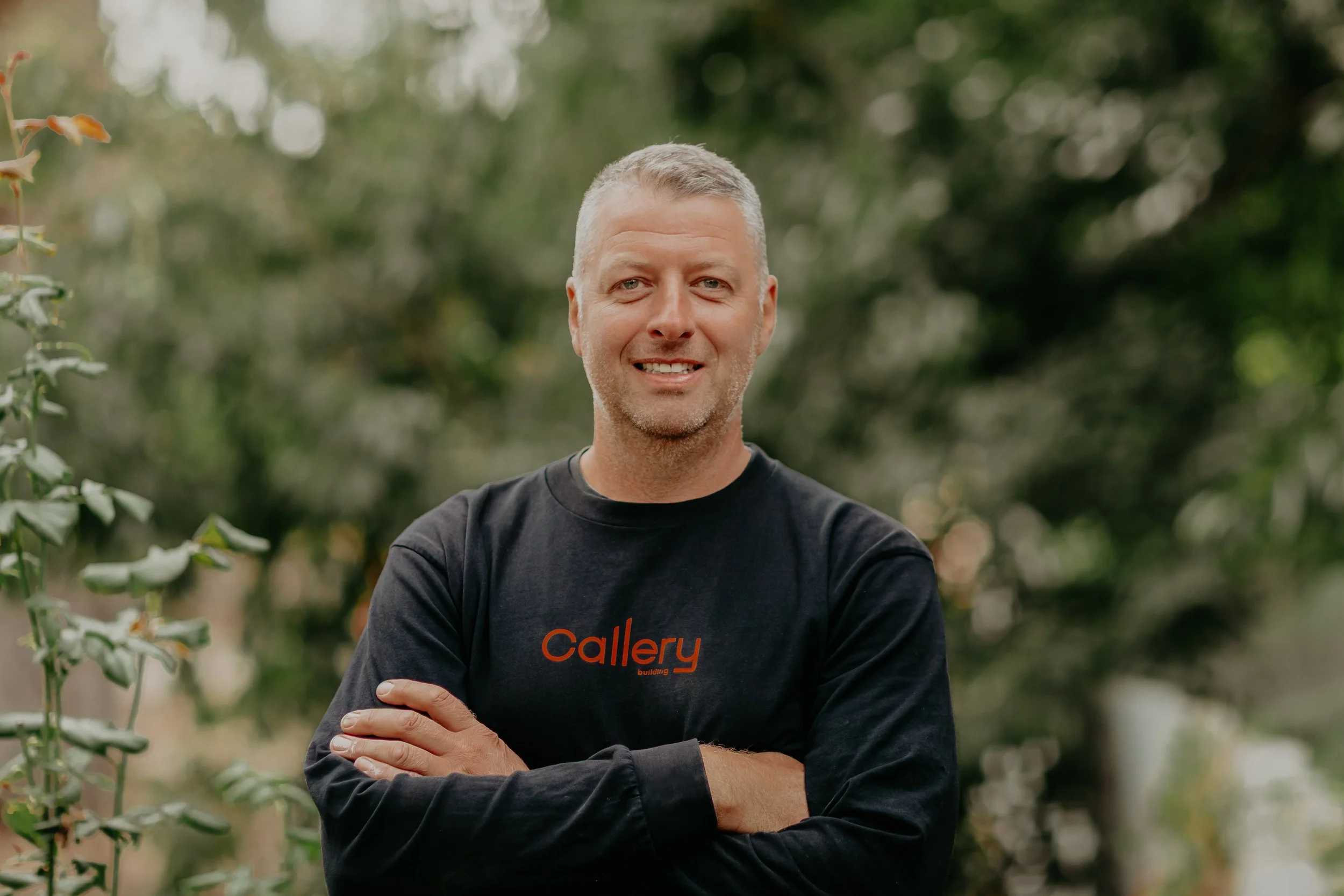 A construction worker inspecting wooden planks at a building site, wearing earmuffs with the brand 'Gallery,' a hat, and safety gear.