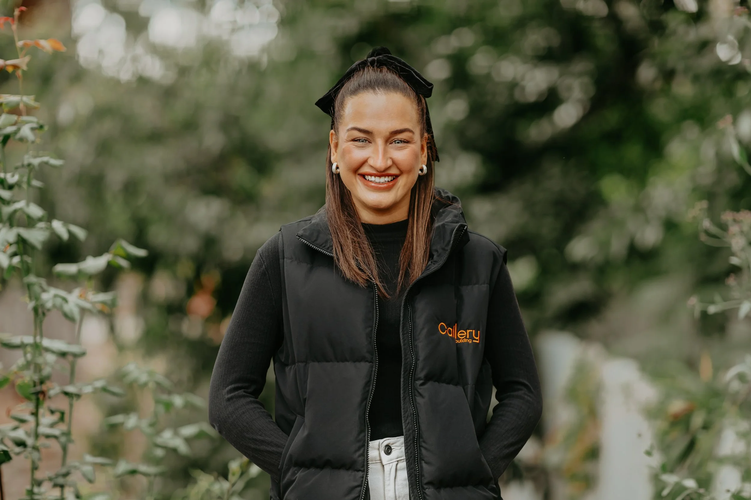 Smiling woman with brown hair tied back with a black bow, wearing a black vest with orange text, and standing outdoors with blurred trees in the background.
