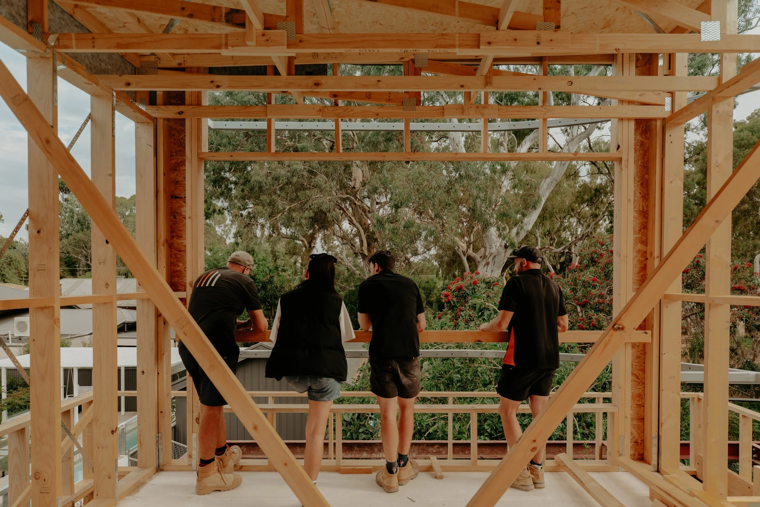 Four people standing inside a wooden framework of a building under construction, looking outdoors at trees and sky.