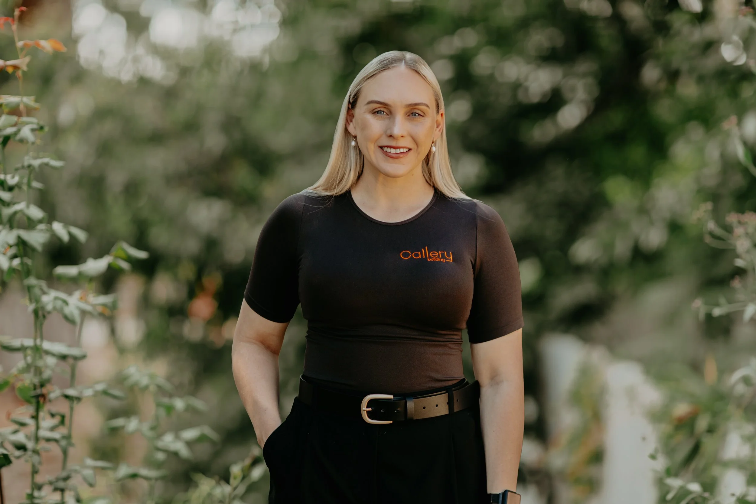 A woman with long blonde hair, wearing a black t-shirt with orange text, black pants with a belt, stands outdoors in front of green foliage, smiling at the camera.