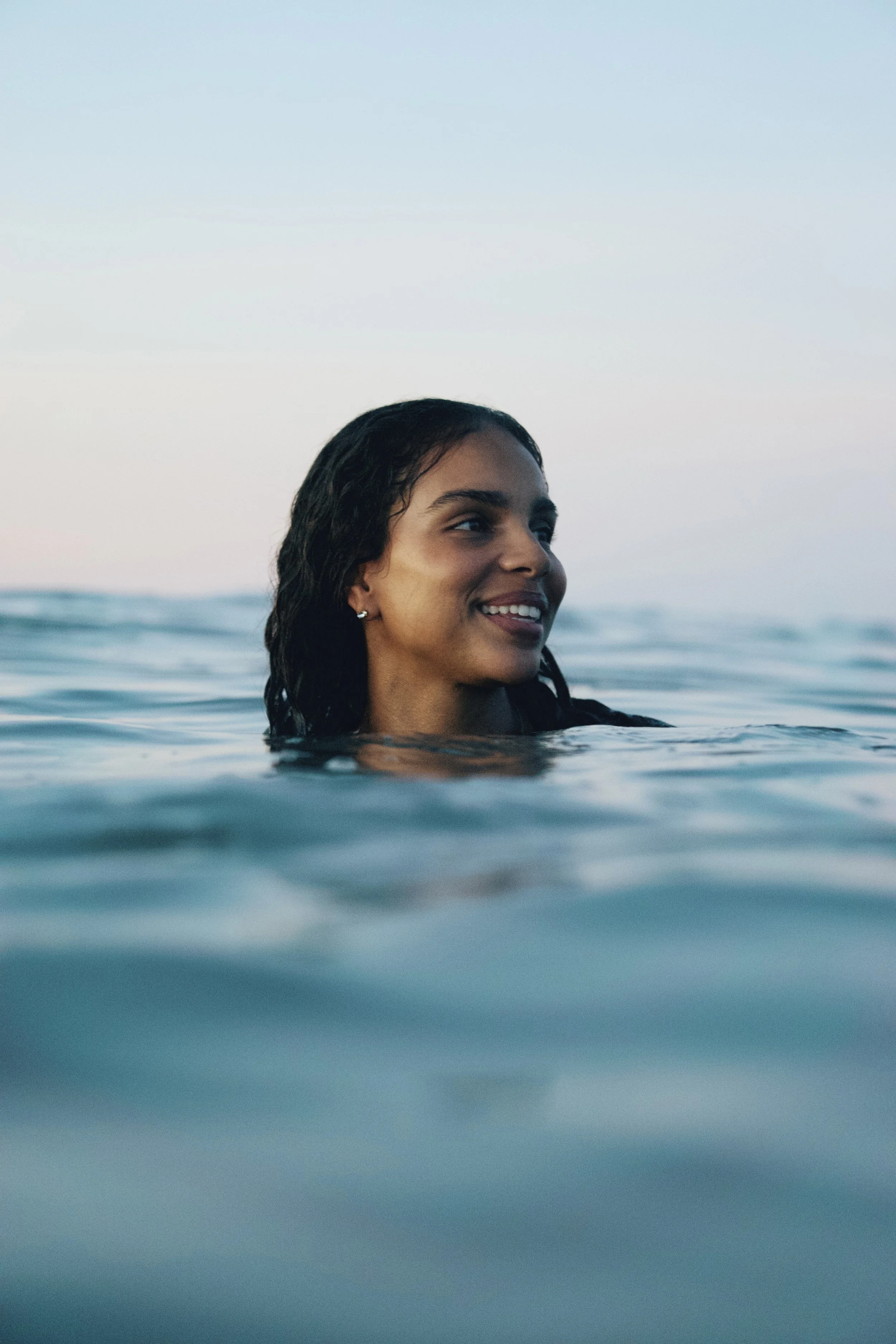Close-up of a smiling woman with wet hair in the ocean, against a pastel sky at dusk.