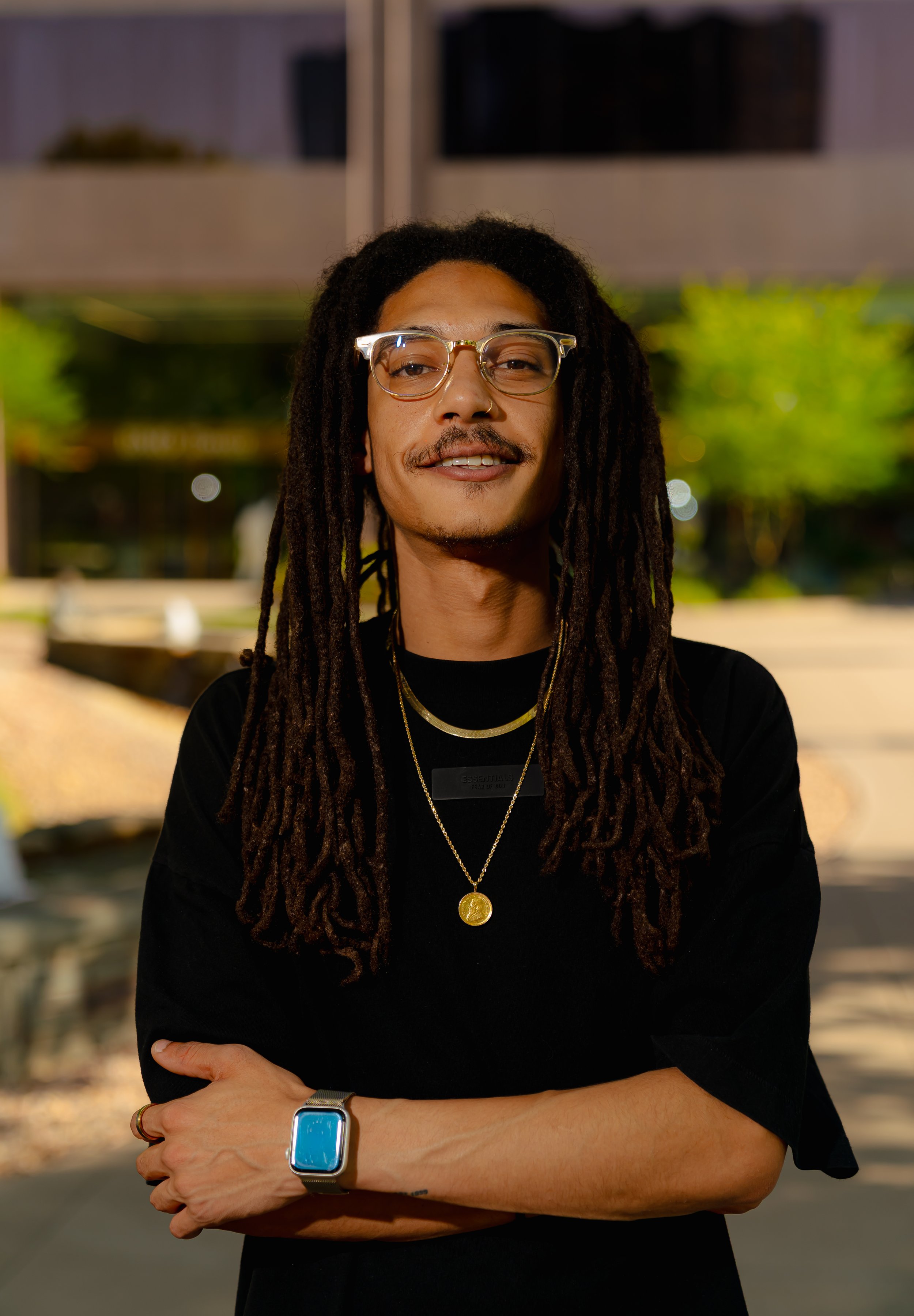 A person with long dreadlocks, glasses, and a mustache, wearing a black t-shirt, gold jewelry, and a smartwatch, standing outside at night with blurred background lights and greenery.