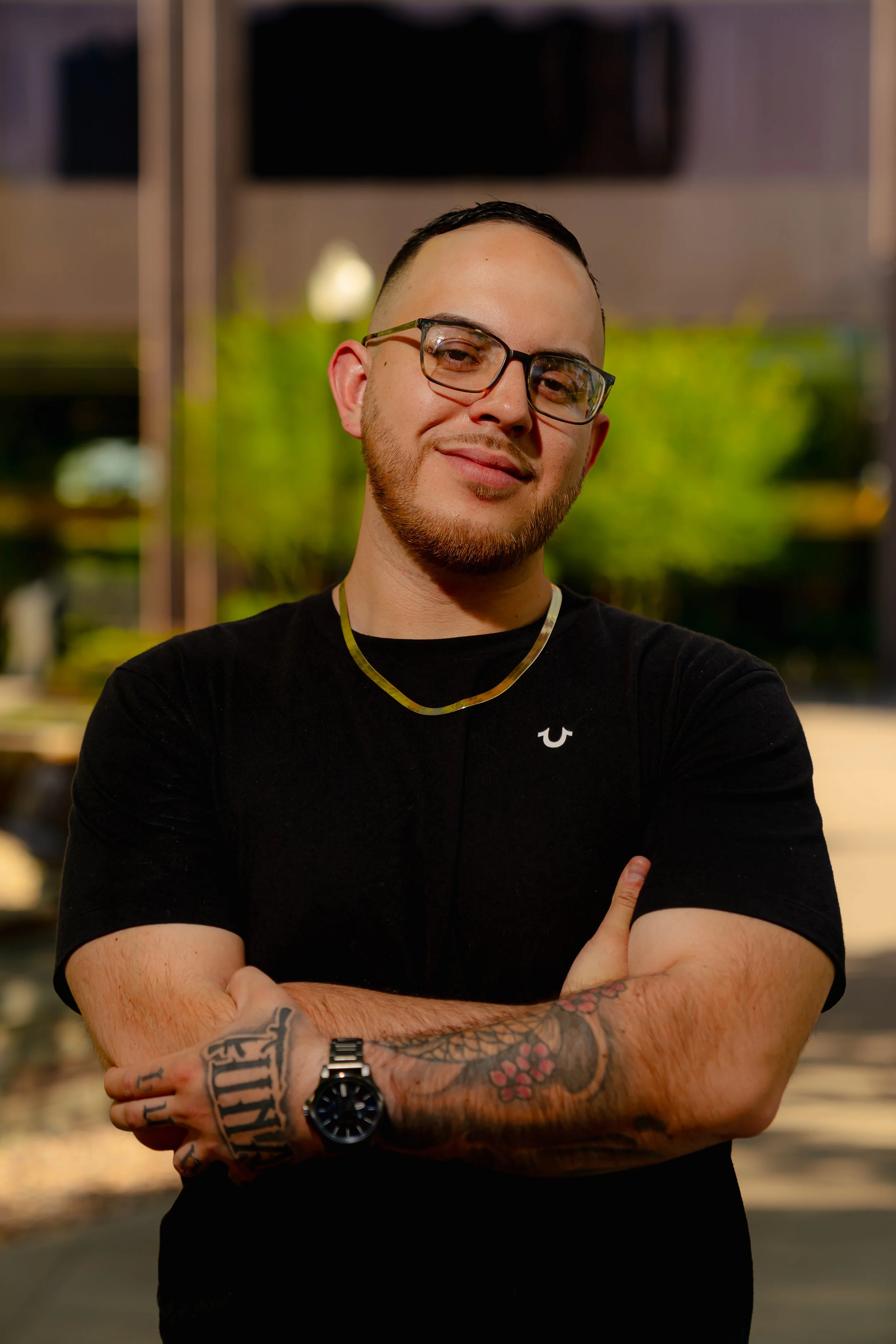 A young man with glasses, facial hair, and tattoos on his arms, standing outdoors with arms crossed and smiling.