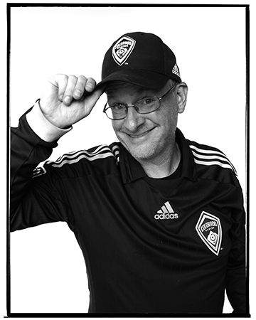 A man in a black adidas jacket and cap, smiling, tipping his cap. The cap and jacket feature a sports team logo.
