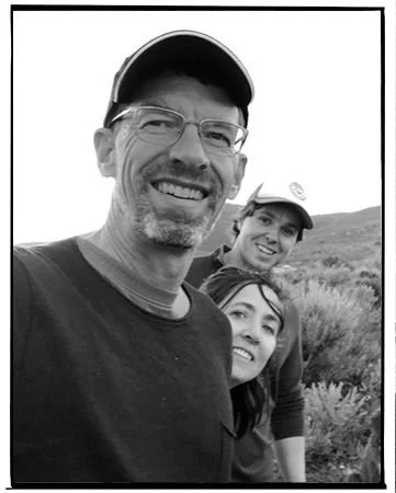 Black-and-white photo of three smiling people outdoors, with hills and vegetation in the background. A man wearing glasses and a baseball cap is in the foreground, with a woman and a girl behind him, all enjoying nature.