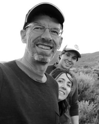 A black-and-white photo of three people outdoors, smiling and wearing hats. The man in the front wears glasses and a baseball cap. Behind him are a woman and a child, also wearing caps, with a natural landscape in the background.