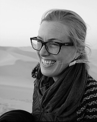 Smiling woman with curly hair, glasses, hoop earrings, layered necklaces, wearing a striped shirt, in an indoor setting.