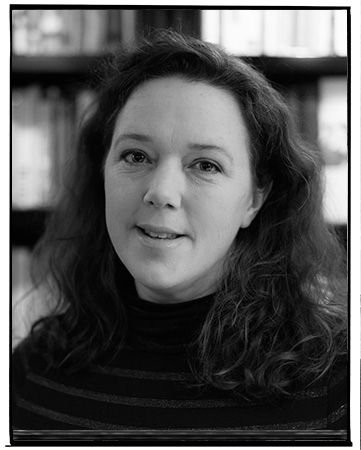Black and white portrait of a woman with shoulder-length wavy hair, wearing a striped turtleneck sweater, inside a room with bookshelves in the background.